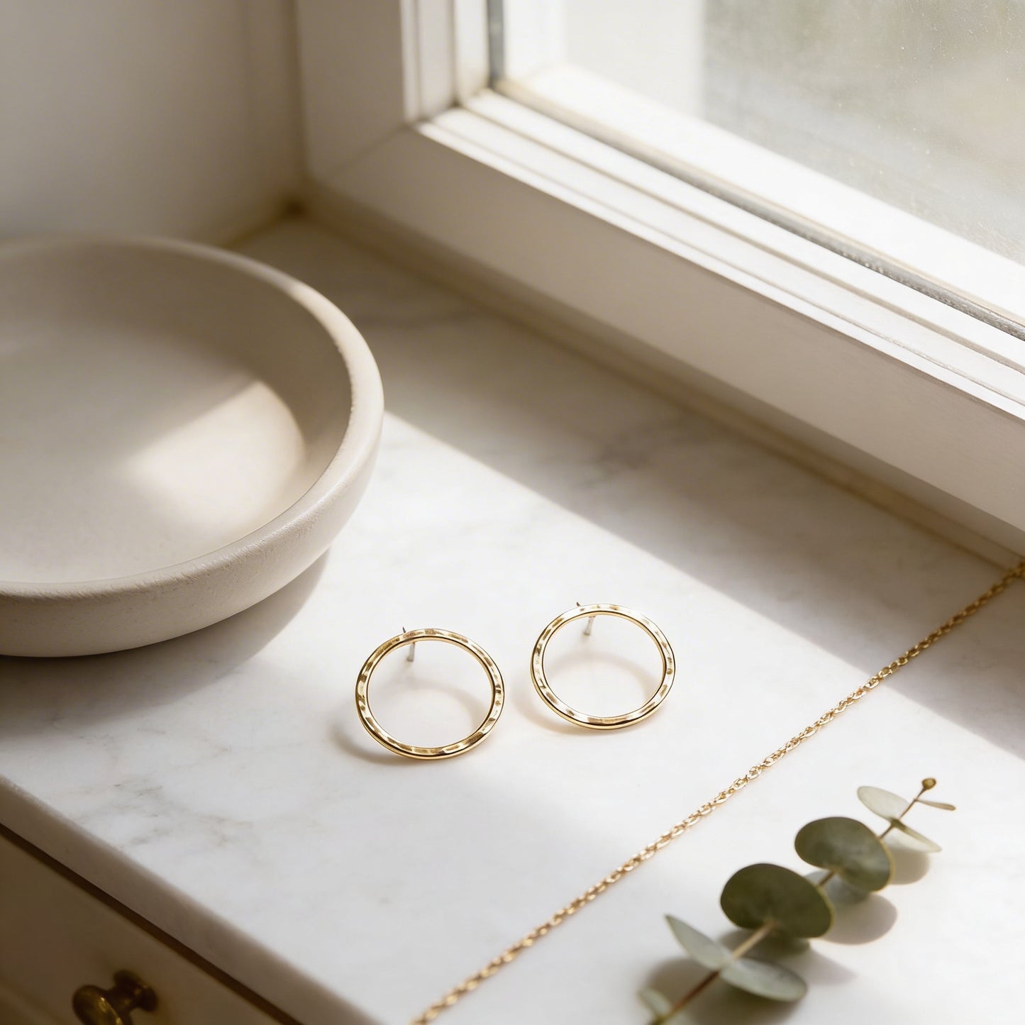 a pair of gold hoop earrings with a white bowl and a green plant on a white marble countertop.