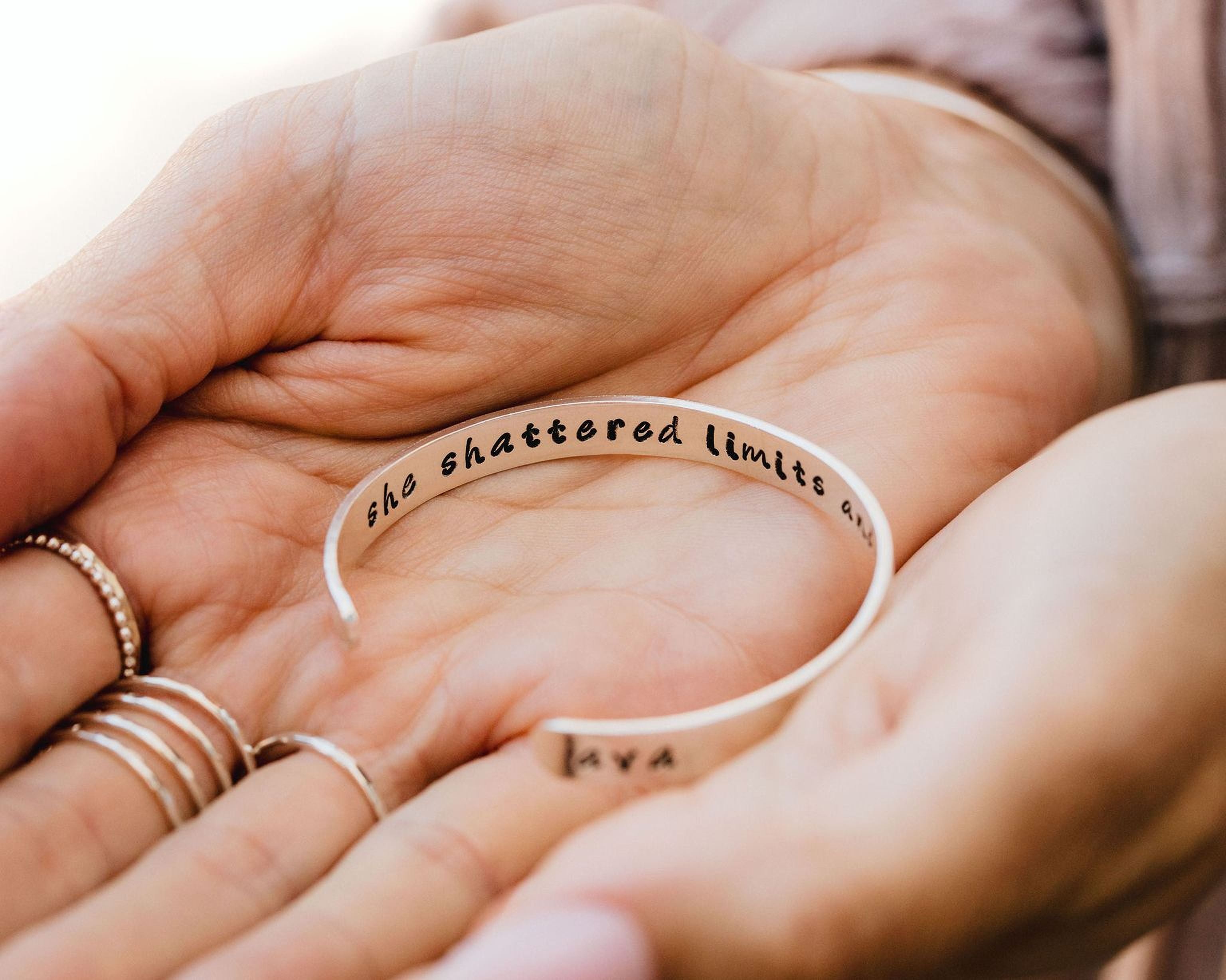 A person's hand holding a bracelet with the text "she shattered limits" engraved on it.