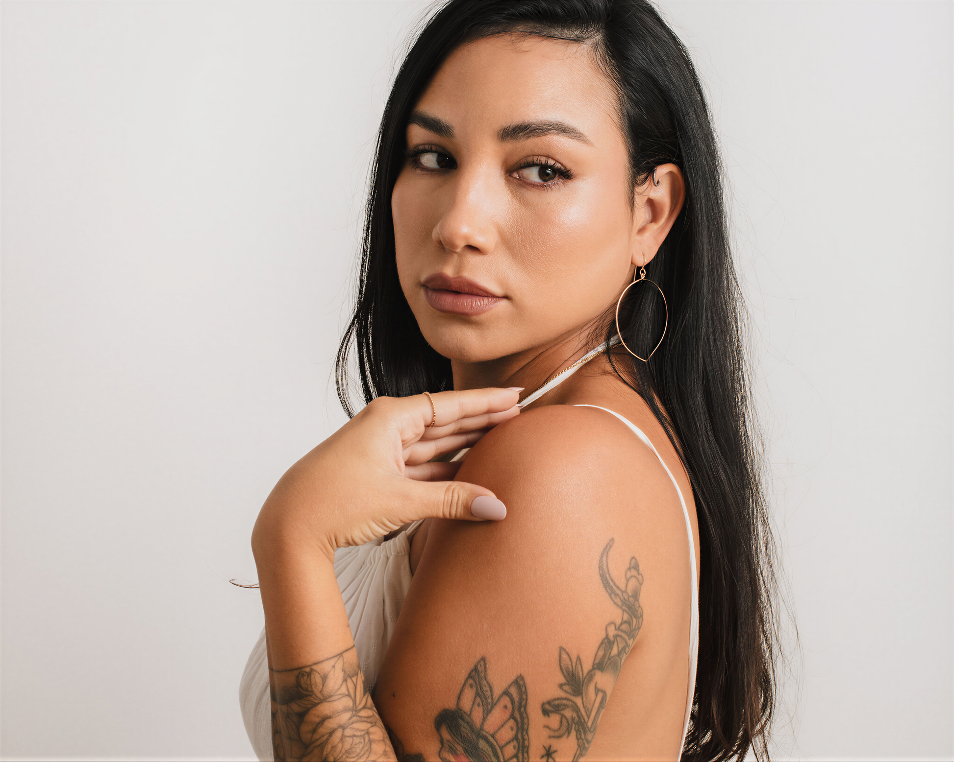 A woman with long dark hair and tattoos on her arm is posing for a portrait against a plain white background.