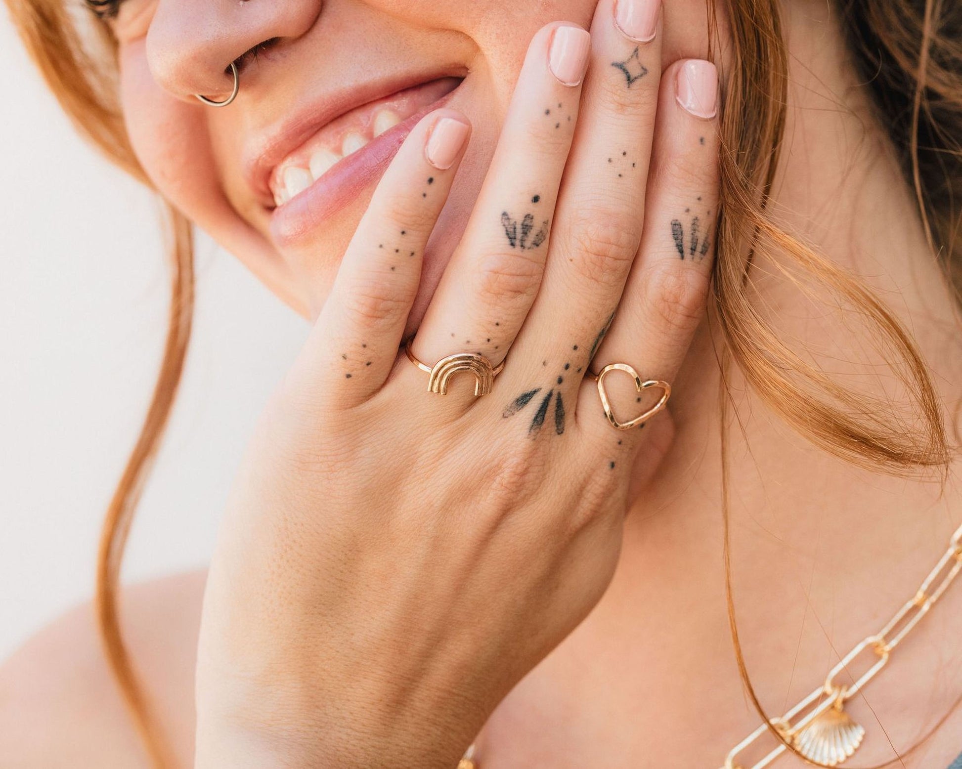 A close-up of a person's hand with multiple rings and tattoos on the fingers, smiling and looking at the camera.
