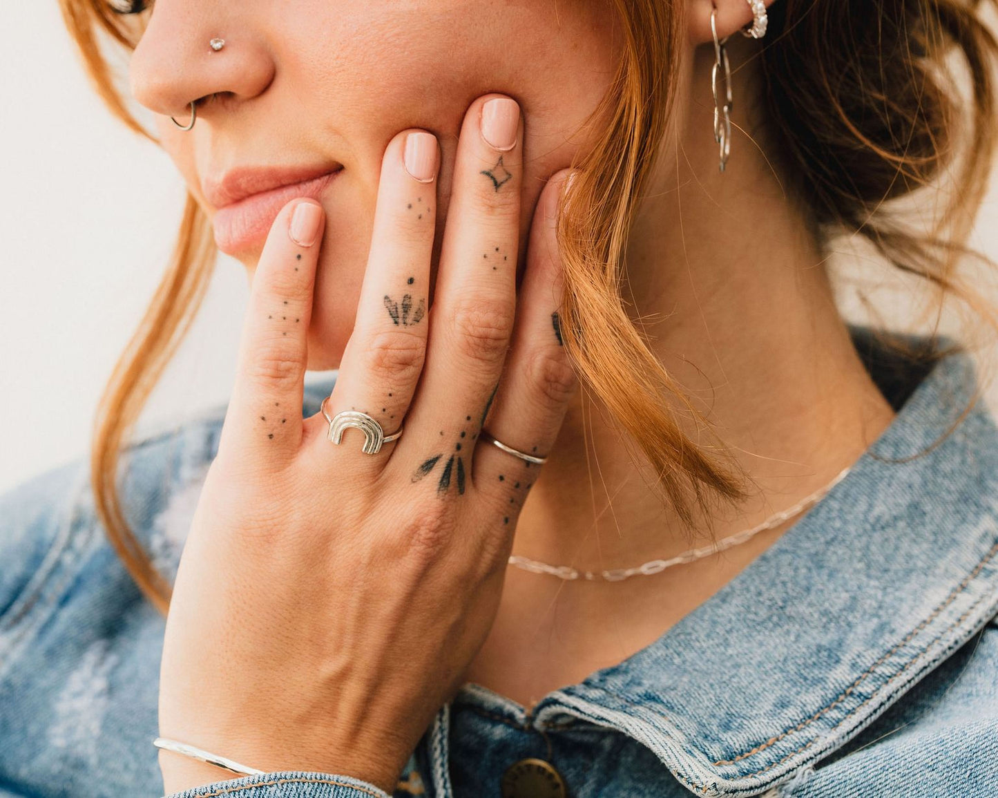 A close-up of a person's hand with tattoos on the fingers, wearing a denim jacket and smiling.