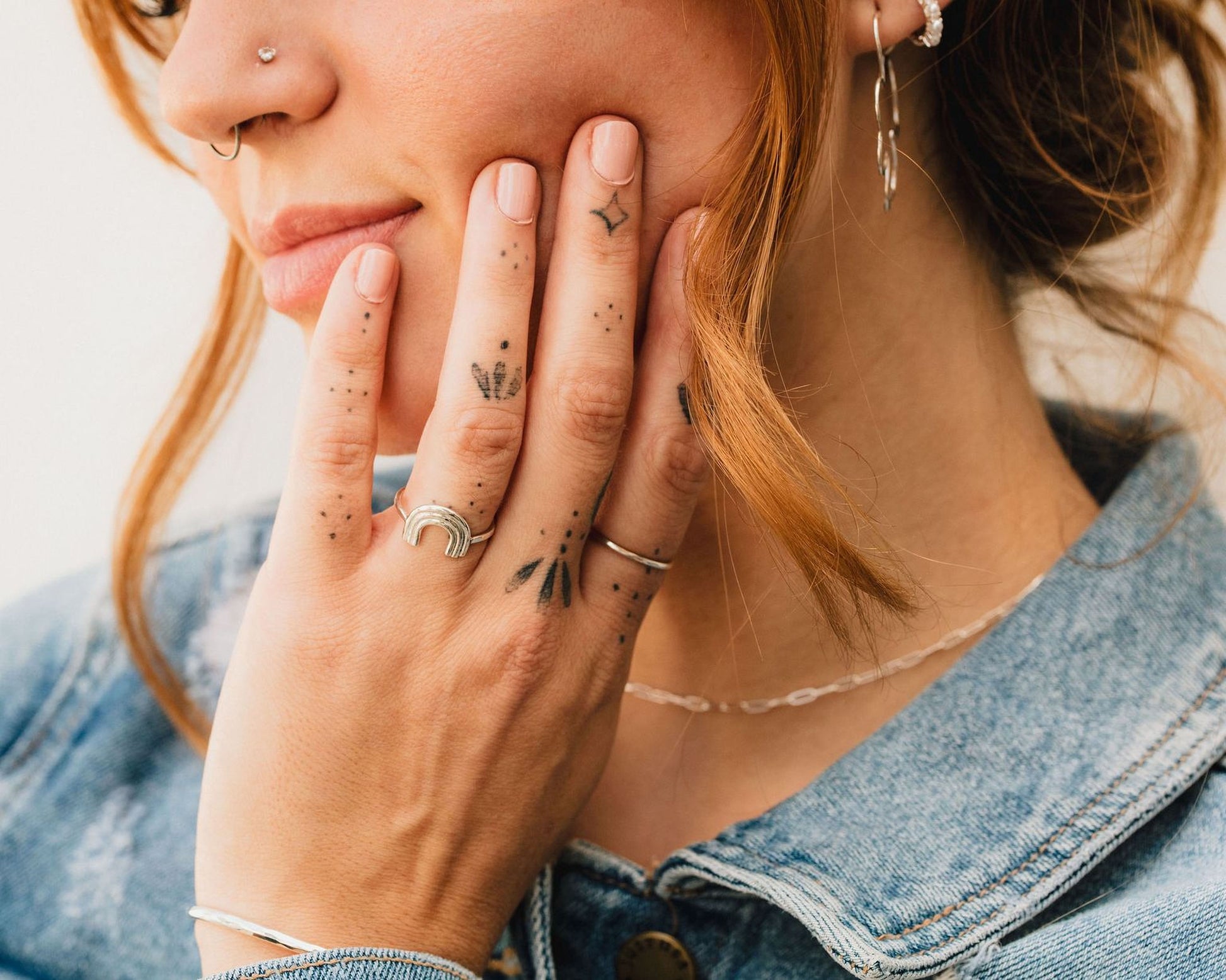 A close-up of a person's hand with tattoos on the fingers, wearing a denim jacket and smiling.