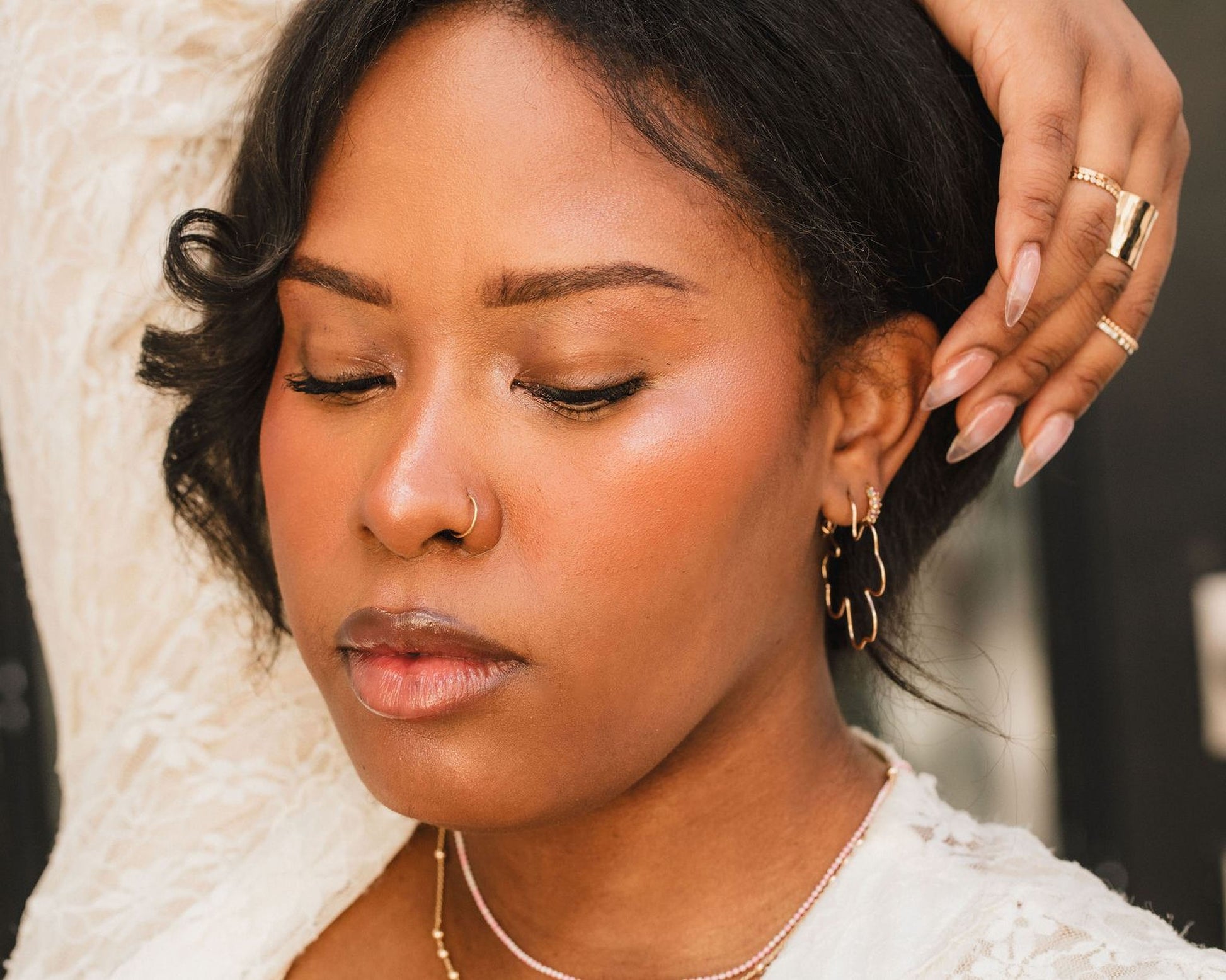A close-up portrait of a woman with her eyes closed, wearing a white top and gold jewelry.