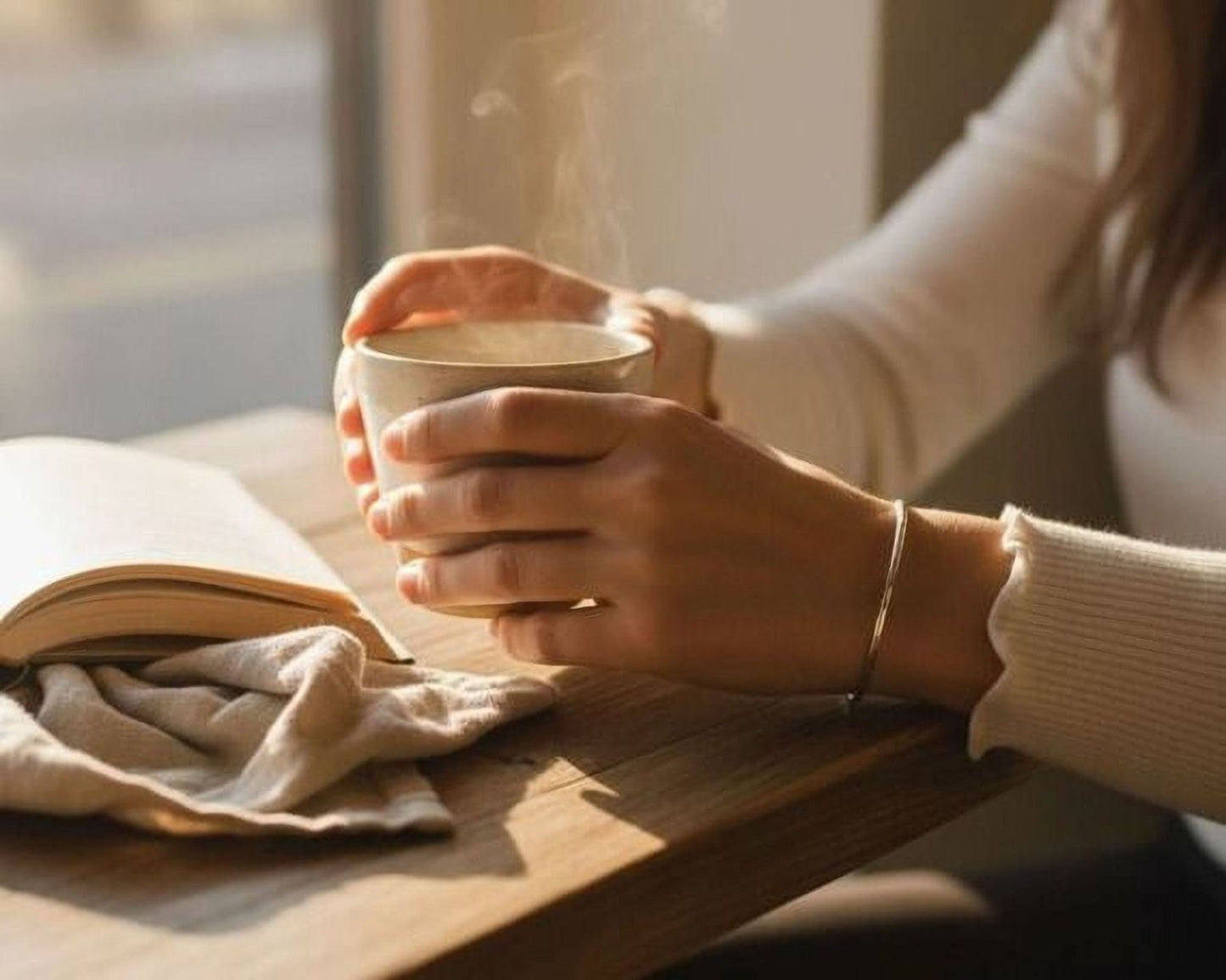 A person's hands holding a steaming cup of coffee while sitting at a wooden table, with an open book nearby.