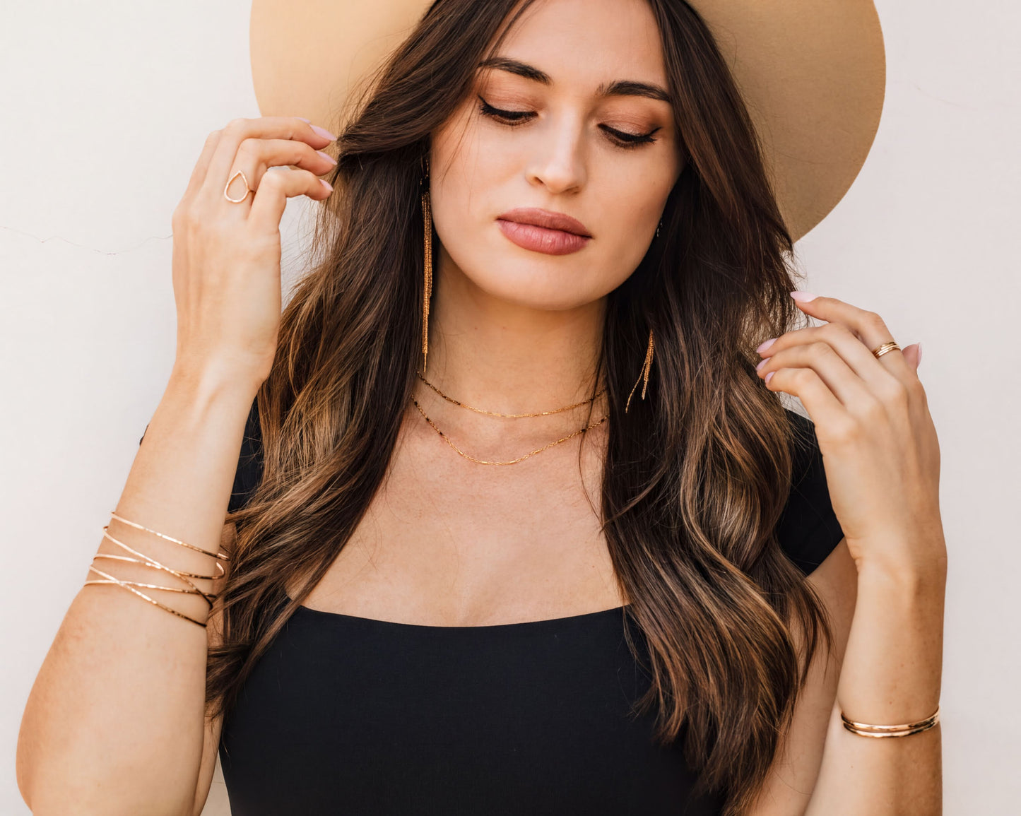 A woman with long brown hair wearing a black top and a wide-brimmed hat, accessorized with multiple rings and bracelets.