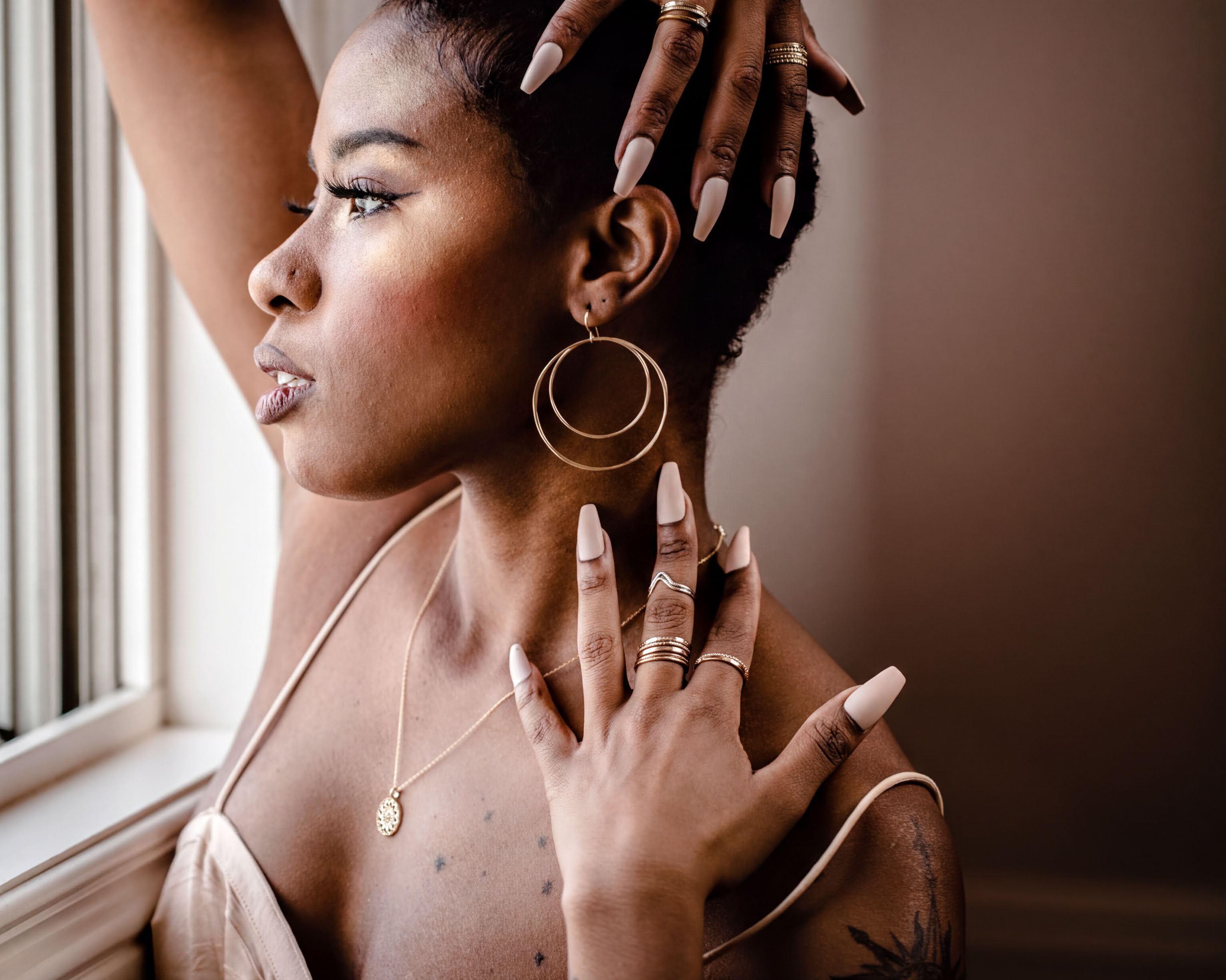 A woman with dark hair and gold jewelry poses with her hands on her head, wearing a beige dress and a necklace.