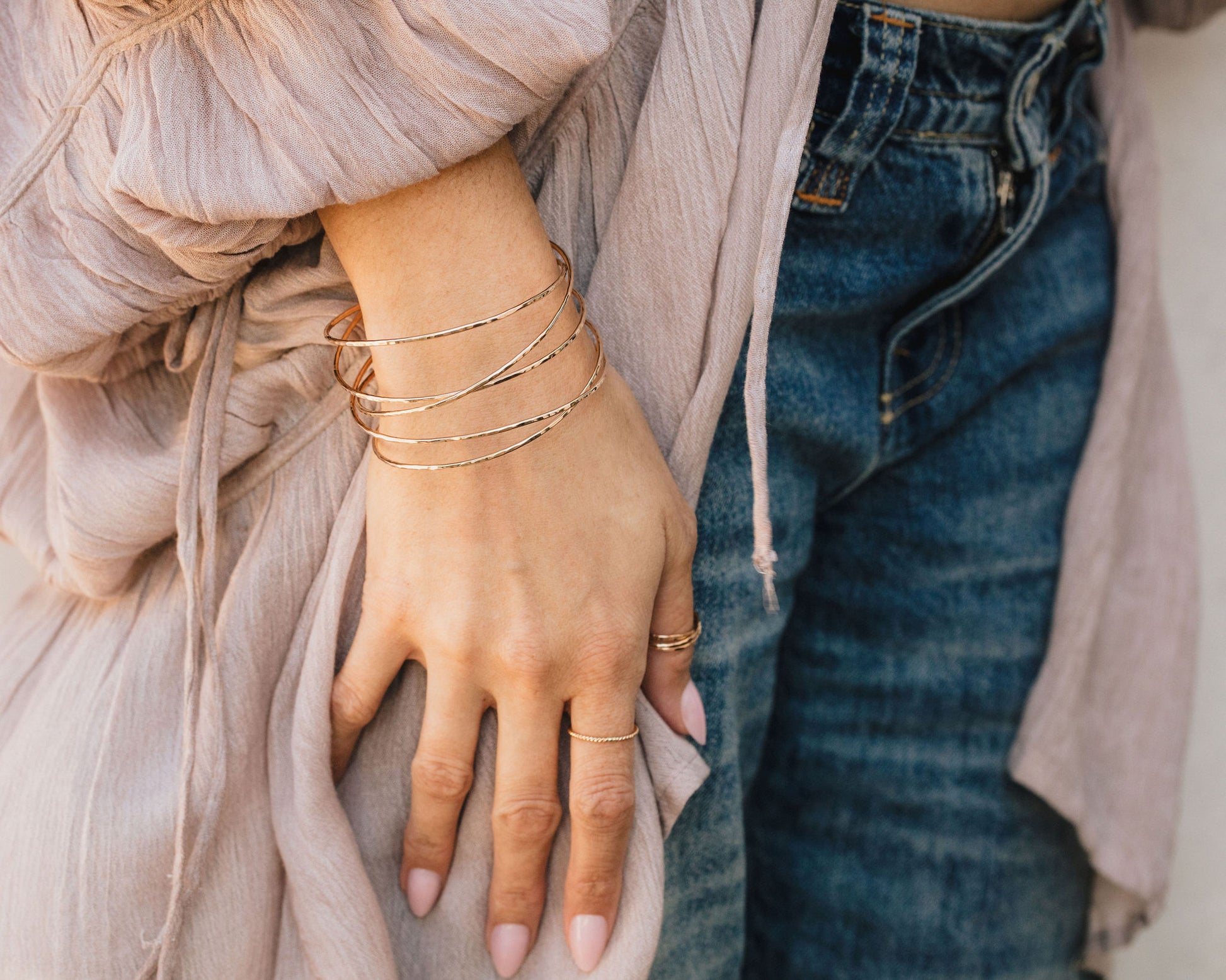 A close-up view of a person's hand wearing multiple gold bracelets and rings, with a light pink robe and blue jeans visible in the background.