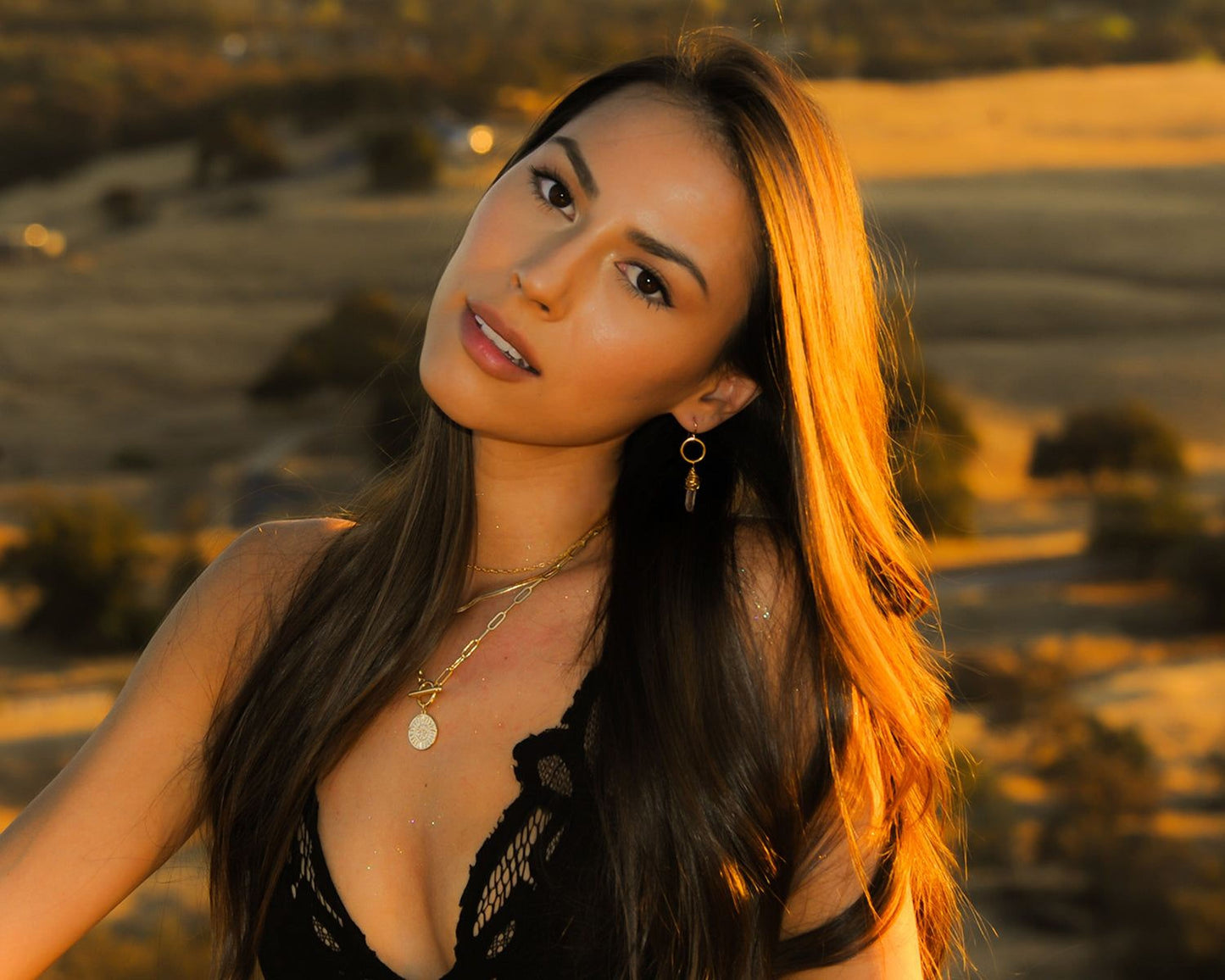 A woman with long dark hair and a black lace top posing in a desert landscape at sunset.