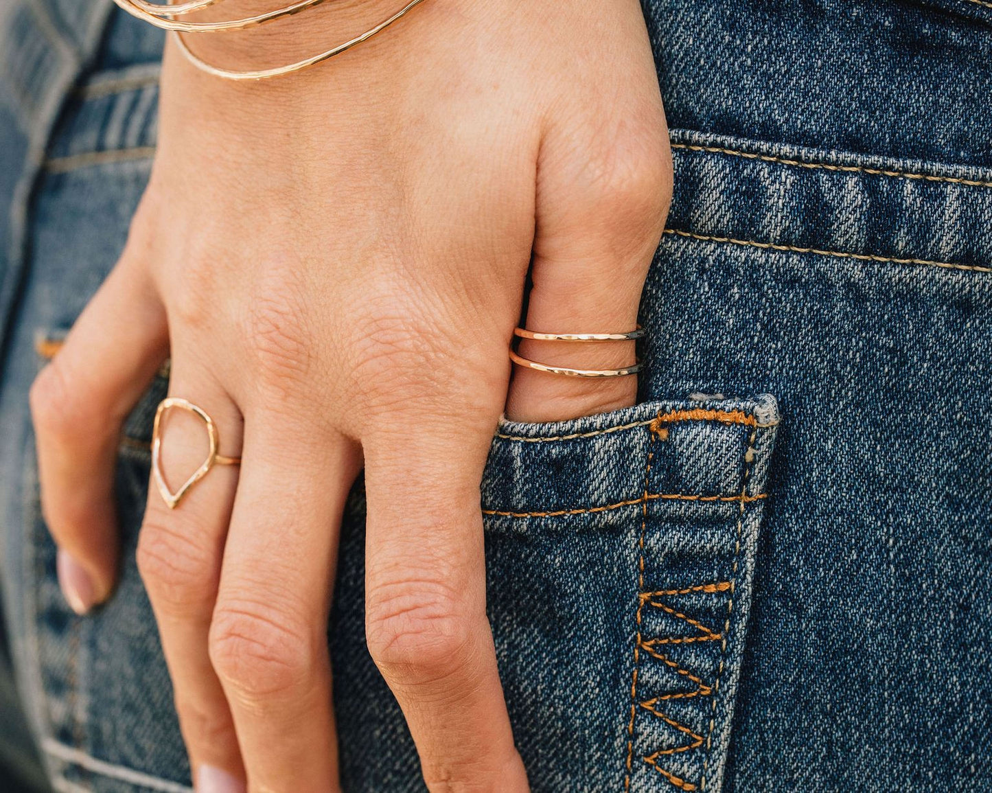 A close-up view of a person's hand wearing multiple rings, with the hand resting on the pocket of a pair of blue jeans.