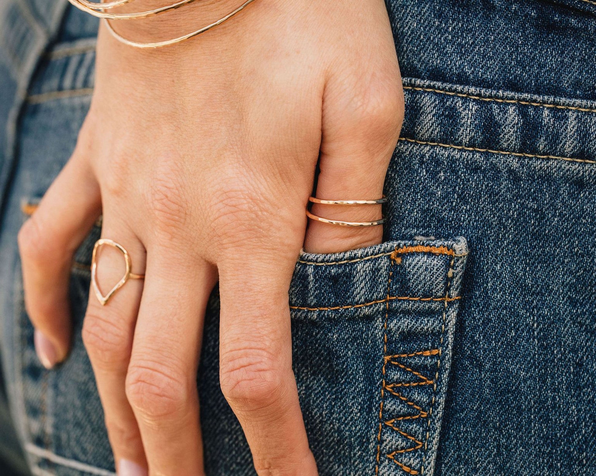 A close-up view of a person's hand wearing multiple rings, with the hand resting on the pocket of a pair of blue jeans.
