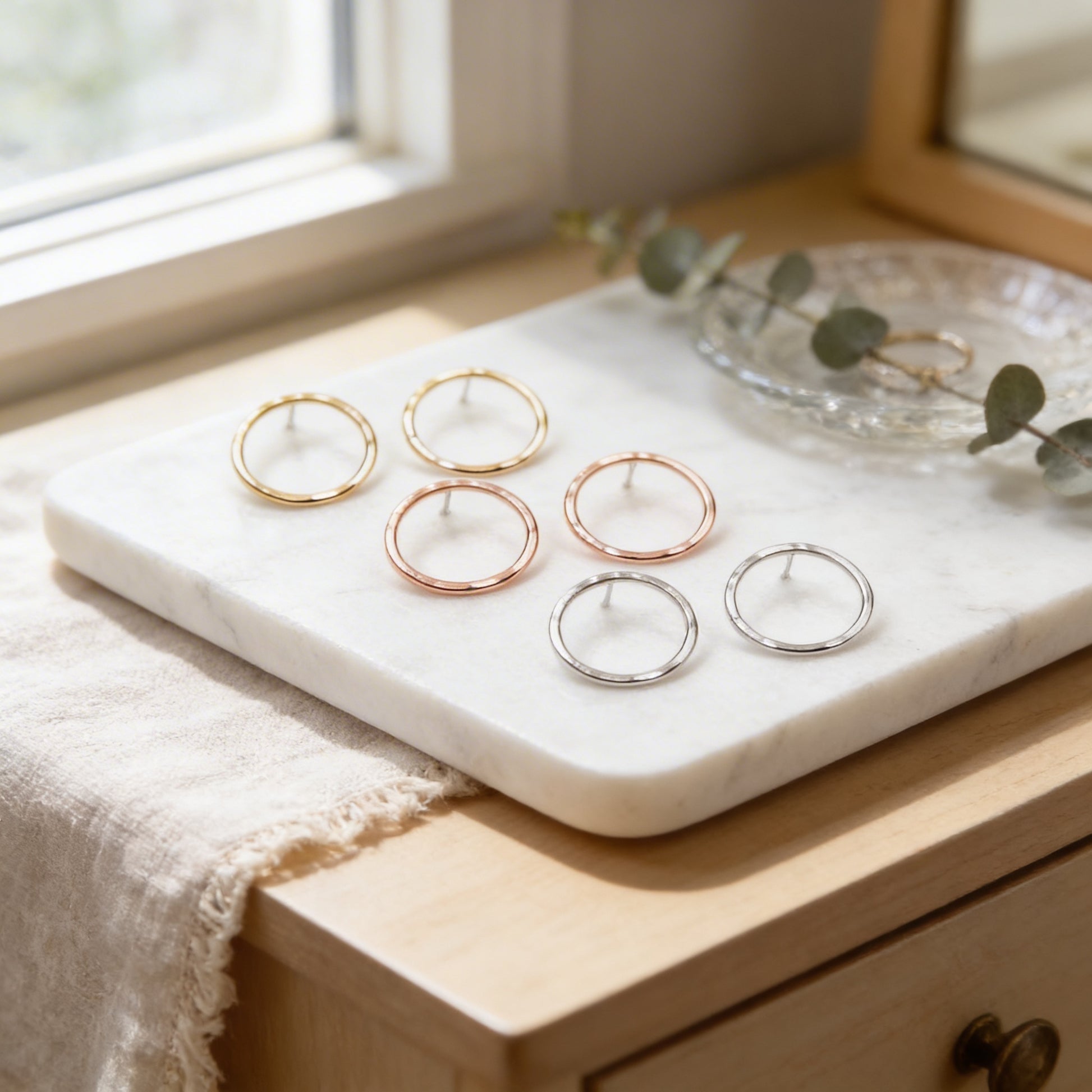 A marble tray displaying a pair of hoop earrings in various colors, including gold, silver, and copper, alongside a small plant or eucalyptus leaf.