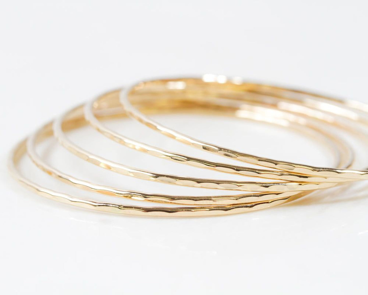 Image shows close up of our ultra thin 14 karat gold bangles. Each one is hand hammered for a light texture. Order one or more in fine 14 karat yellow gold filled. Elevate your stack. Beautiful, polished, shiny. Bracelet width is 1/16 inch wide.