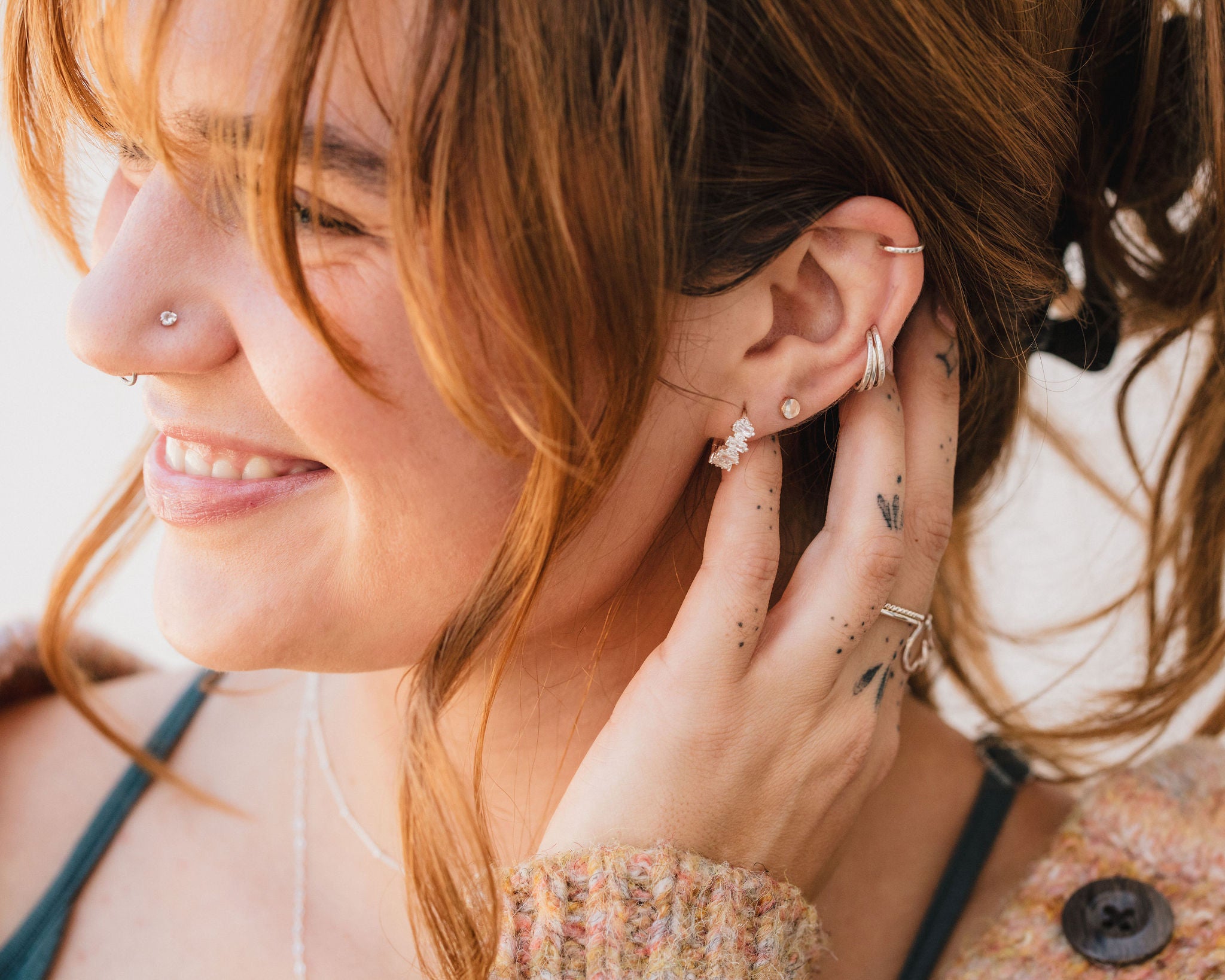 A woman with a nose piercing and multiple earrings is smiling and touching her ear with her hand.