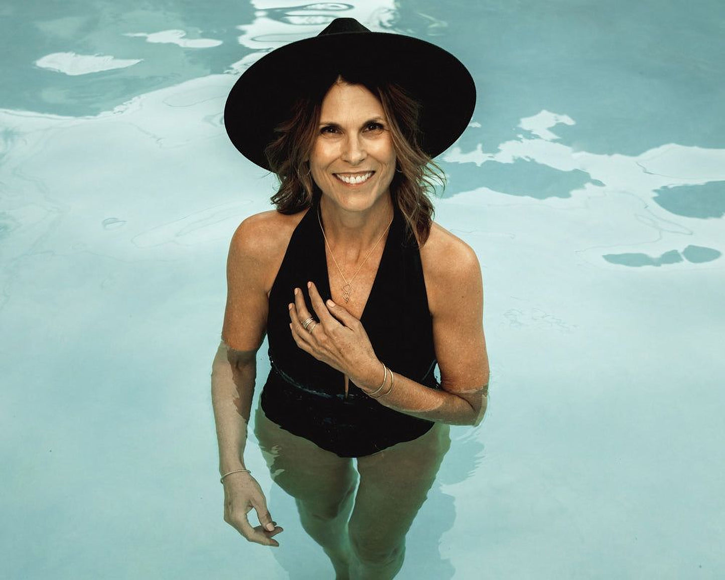 A woman in a black swimsuit and wide-brimmed hat stands in a pool, smiling and looking directly at the camera.