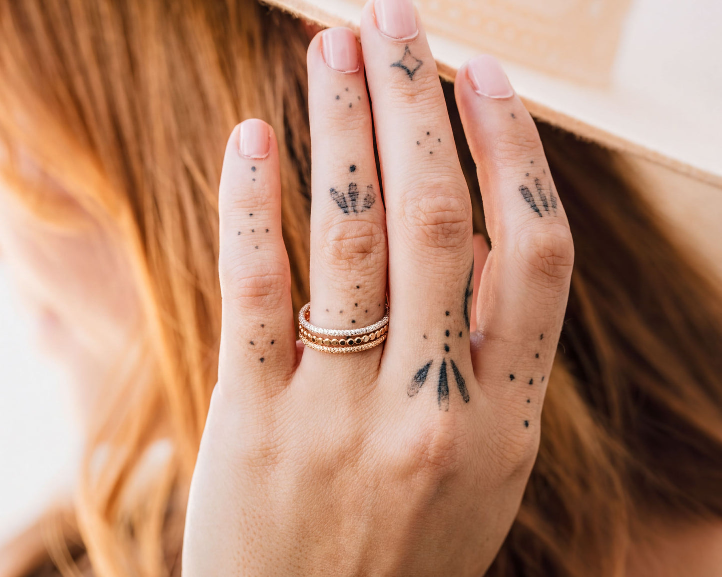 A close-up of a person's hand with tattoos on the fingers, wearing a ring, and partially covering their face.