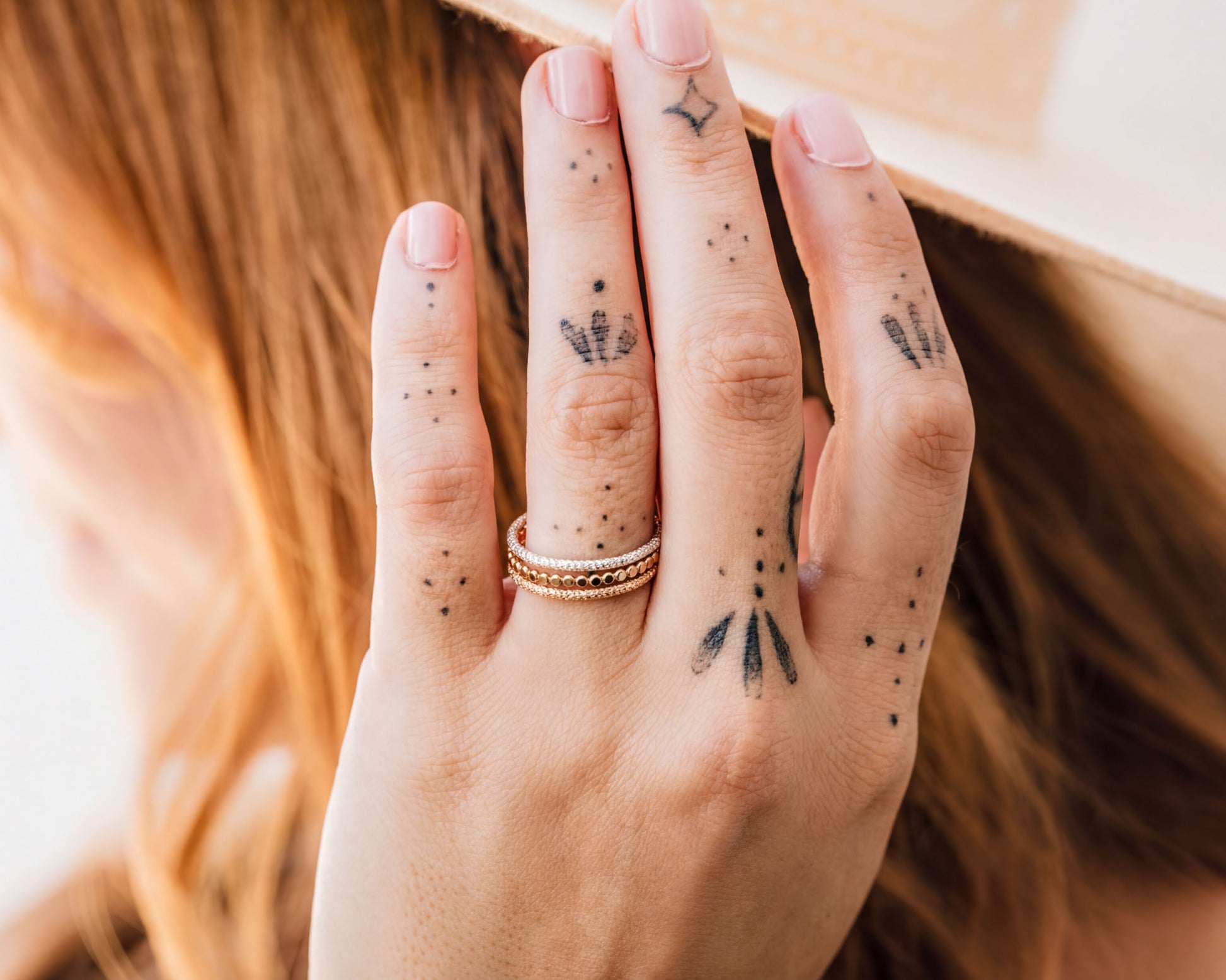 A close-up of a person's hand with tattoos on the fingers, wearing a ring, and partially covering their face.