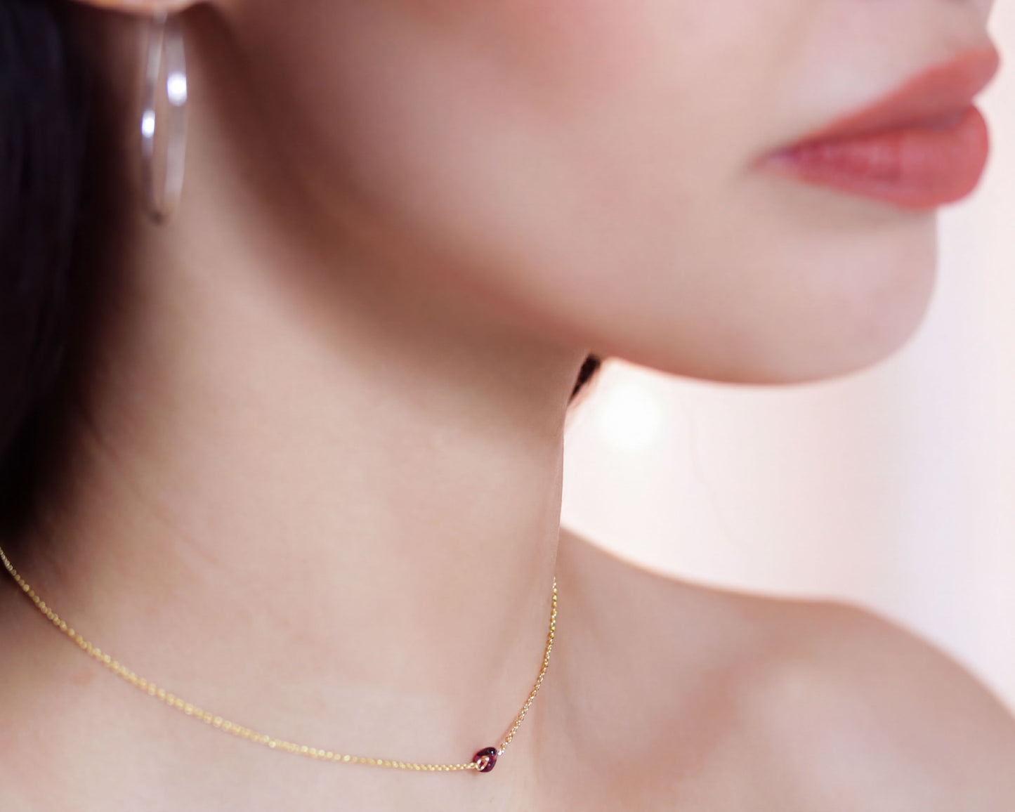 A close-up of a woman's neck and chest area, with a delicate gold necklace and a small red gemstone pendant visible.