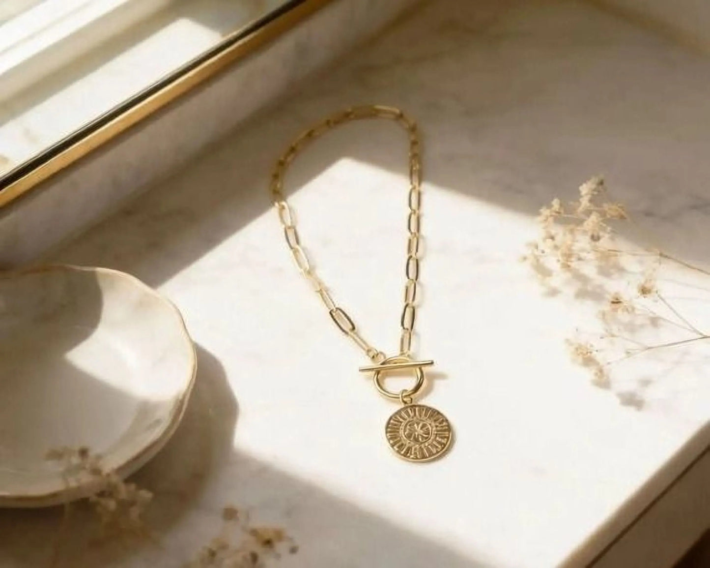 A gold-colored necklace with a pendant is displayed on a marble surface, accompanied by a small white bowl and some dried flowers.