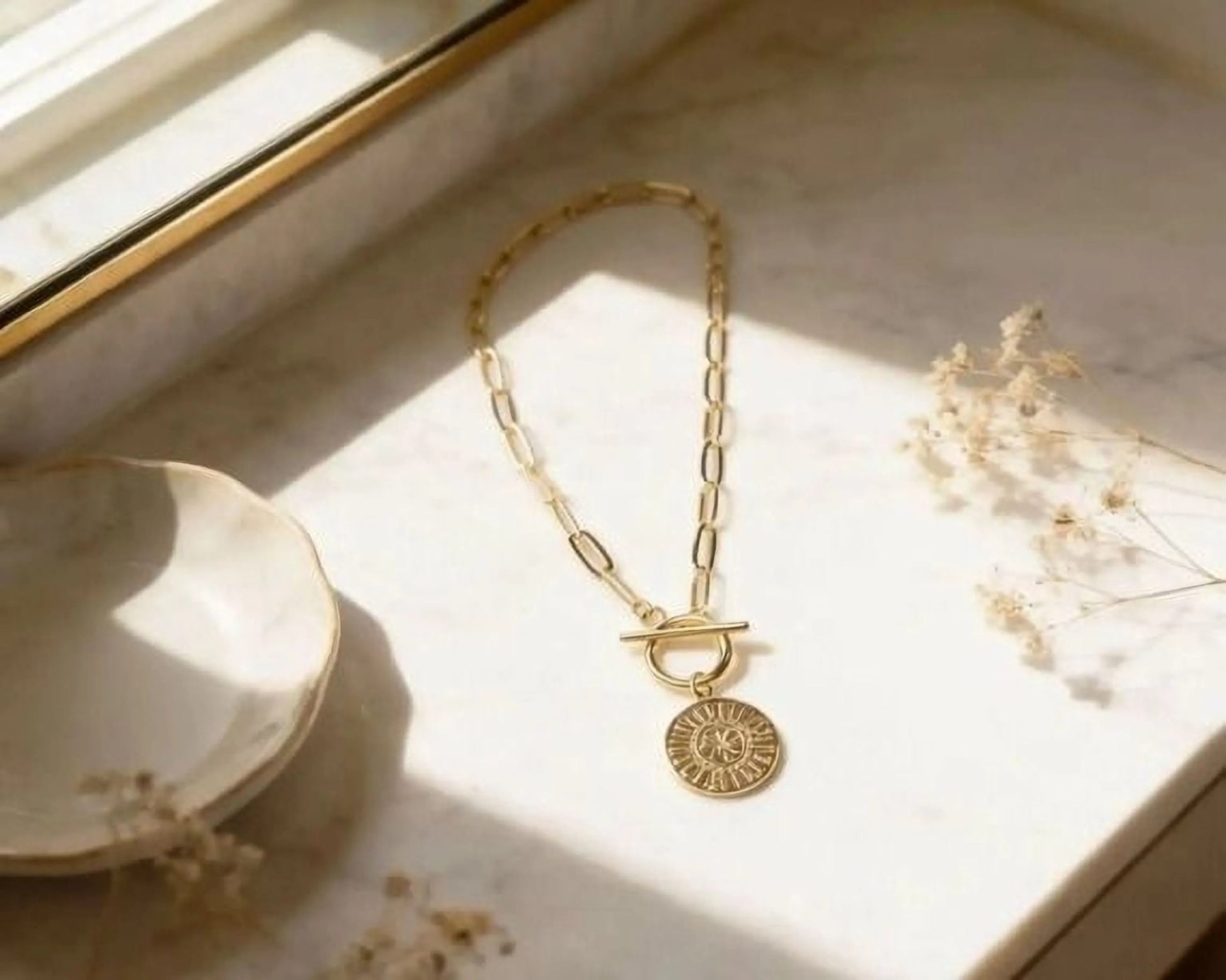 A gold-colored necklace with a pendant is displayed on a marble surface, accompanied by a small white bowl and some dried flowers.