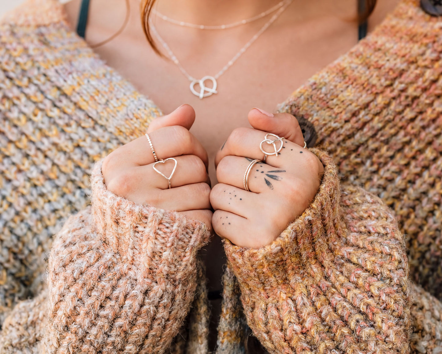 A close-up of a person's hands wearing multiple rings, including heart-shaped and diamond-encrusted rings, against a textured sweater background.