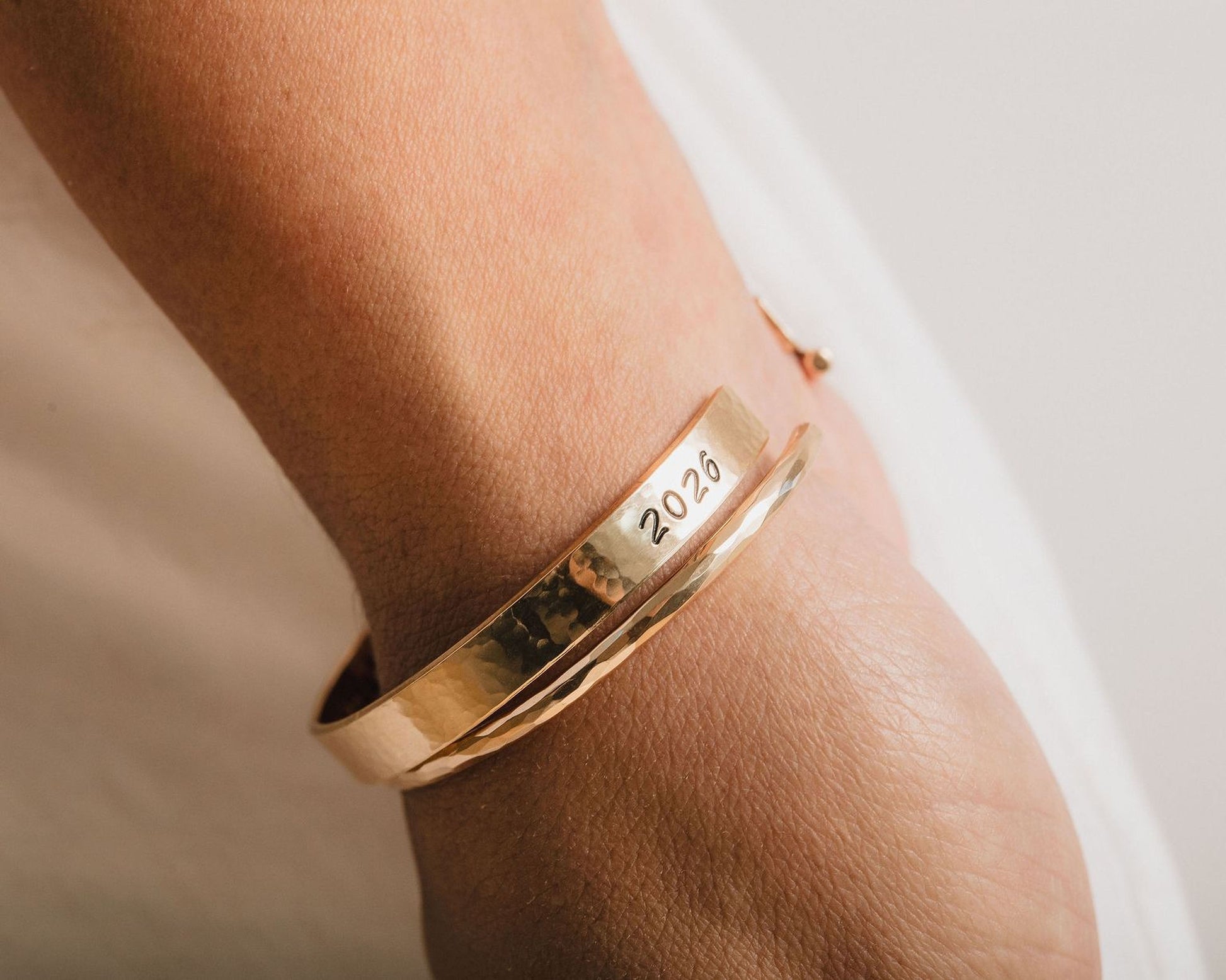 A gold bracelet with the year "2026" engraved on it is worn on a person's wrist.