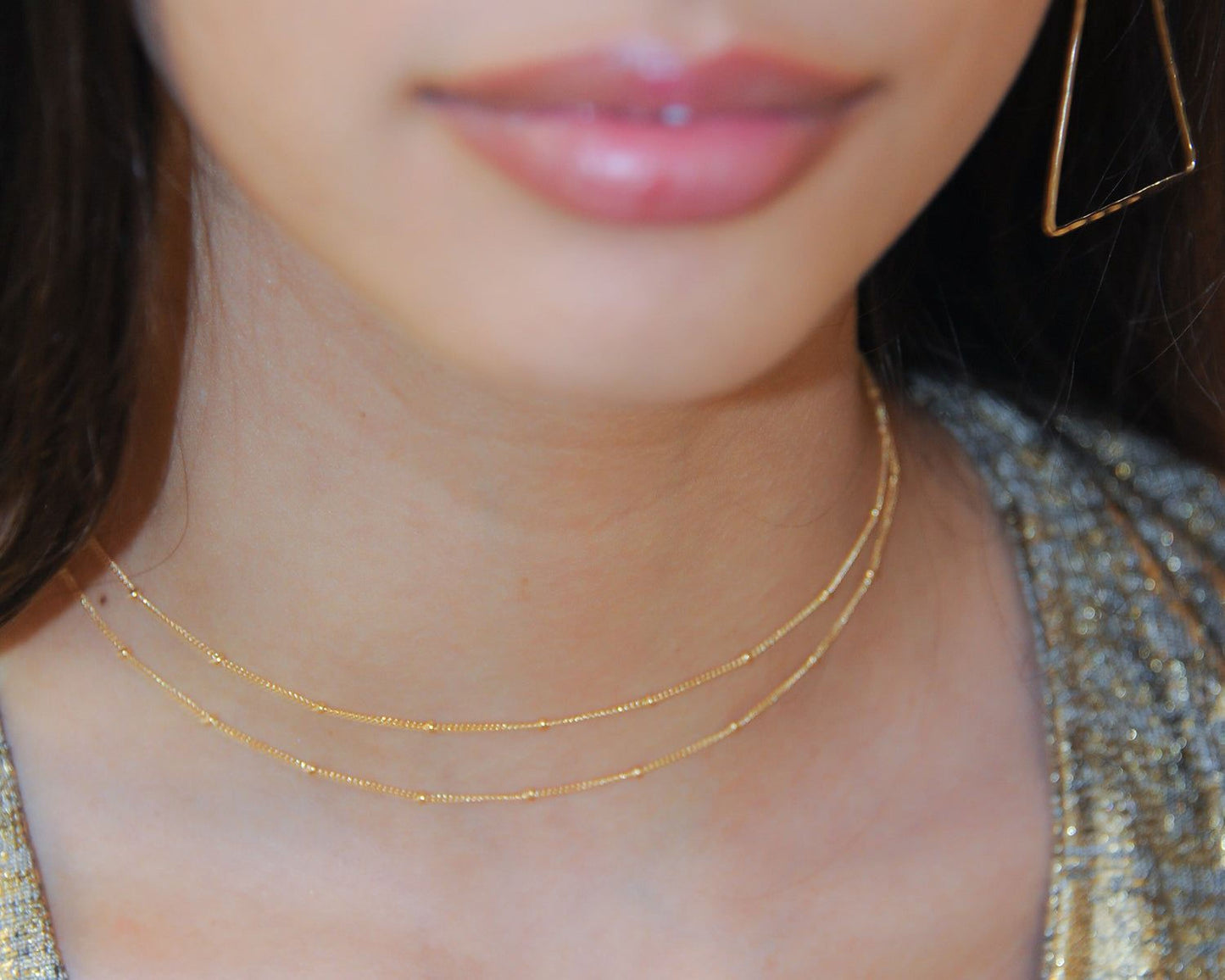 A close-up of a woman's face, focusing on her lips and neck area. She is wearing a gold necklace with two strands.