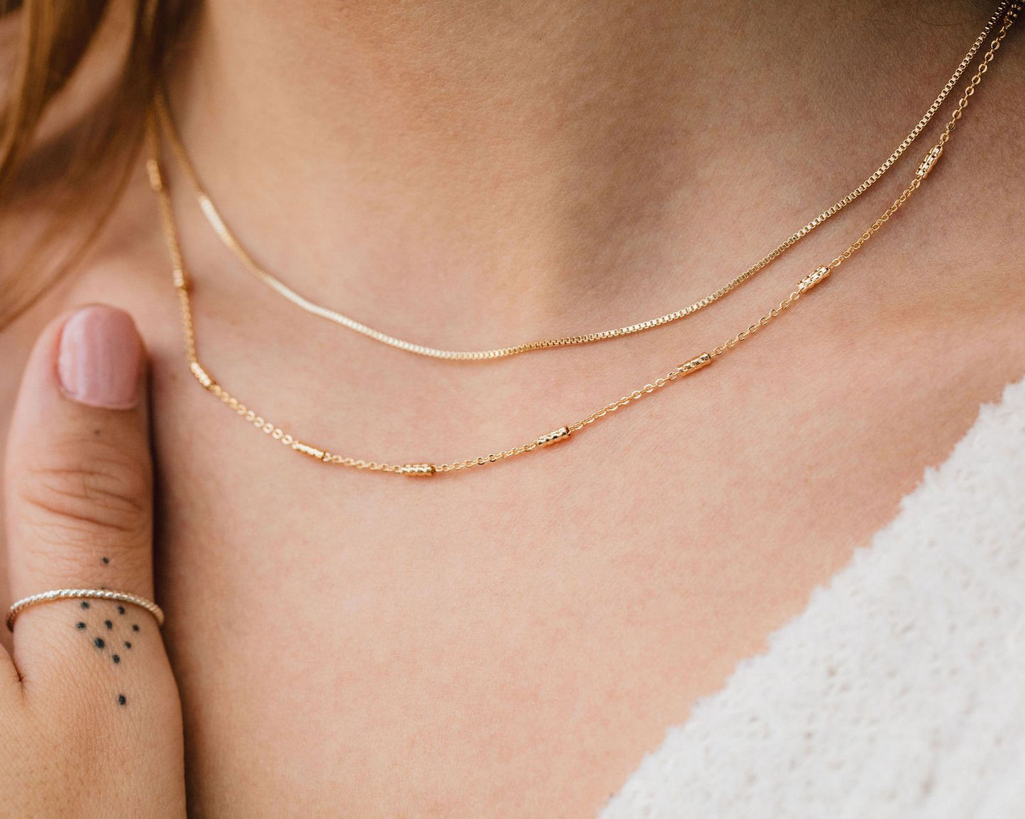 A close-up view of a person's neck and chest area, featuring a delicate gold necklace with multiple strands of chain and a matching ring on the same finger.
