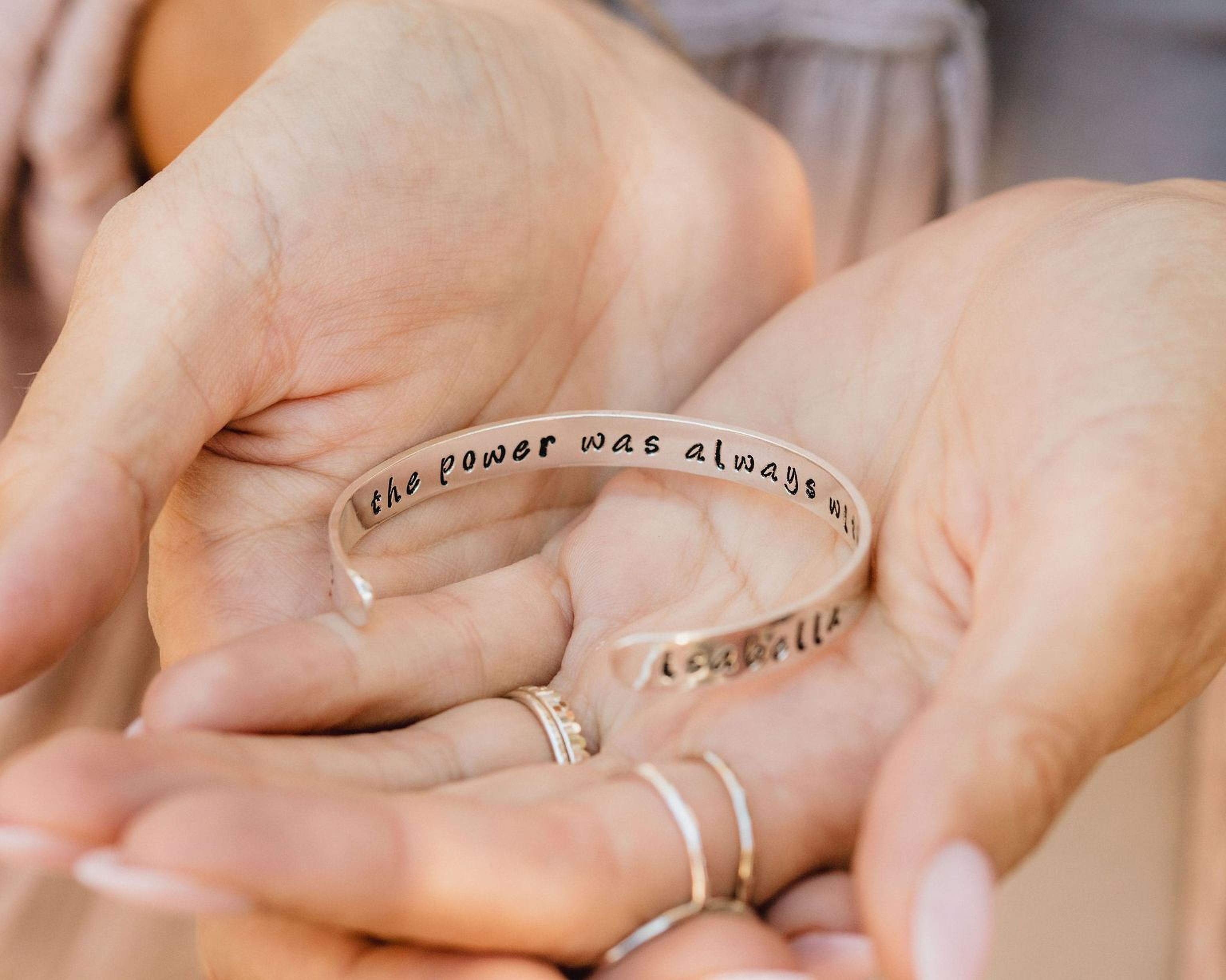 A person's hands holding a silver bracelet with the text "The power was always within" engraved on it.