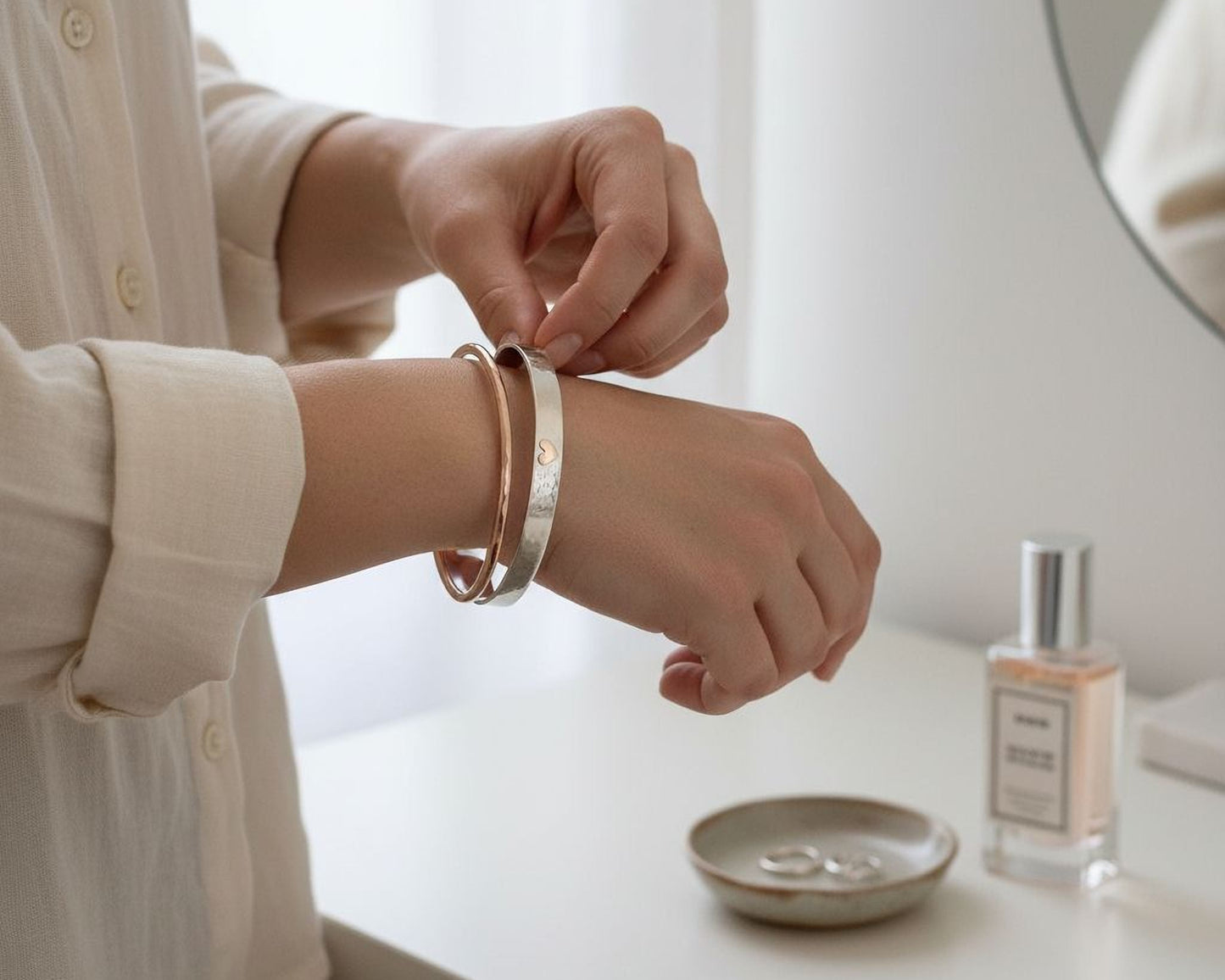 A person is adjusting a bracelet on their wrist while standing near a vanity with a bottle of perfume and a small bowl containing rings.