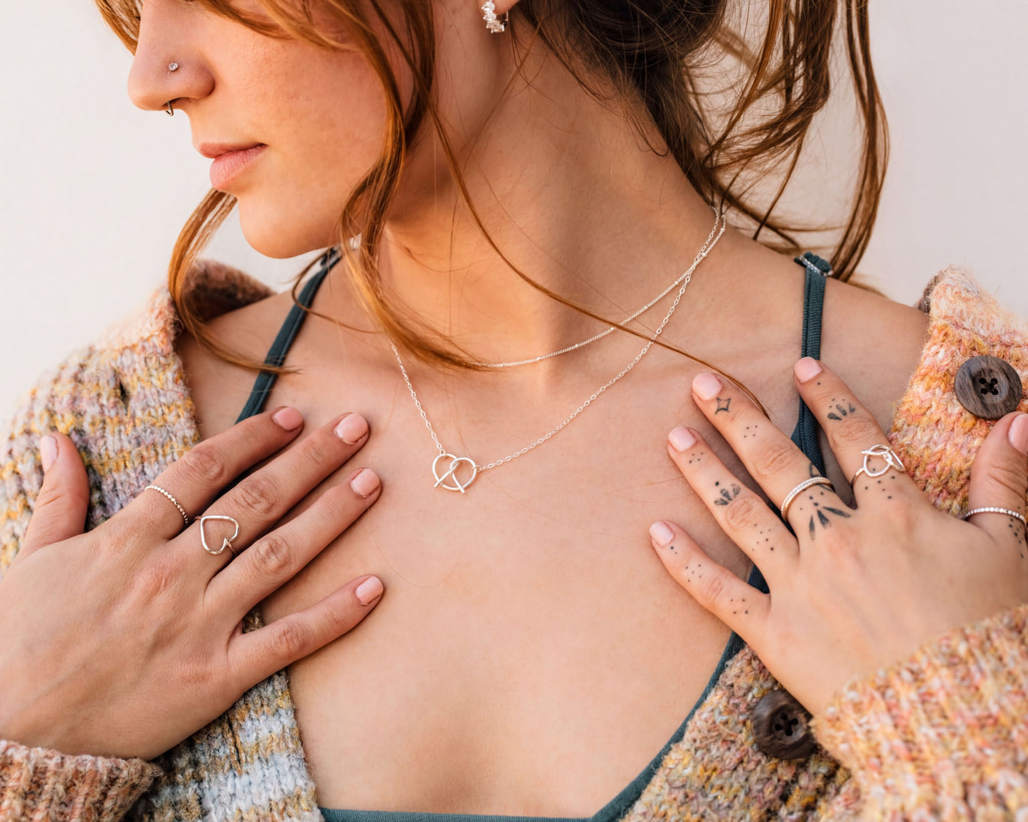 A close-up of a woman's face wearing a necklace and rings, with her hands placed on her chest.