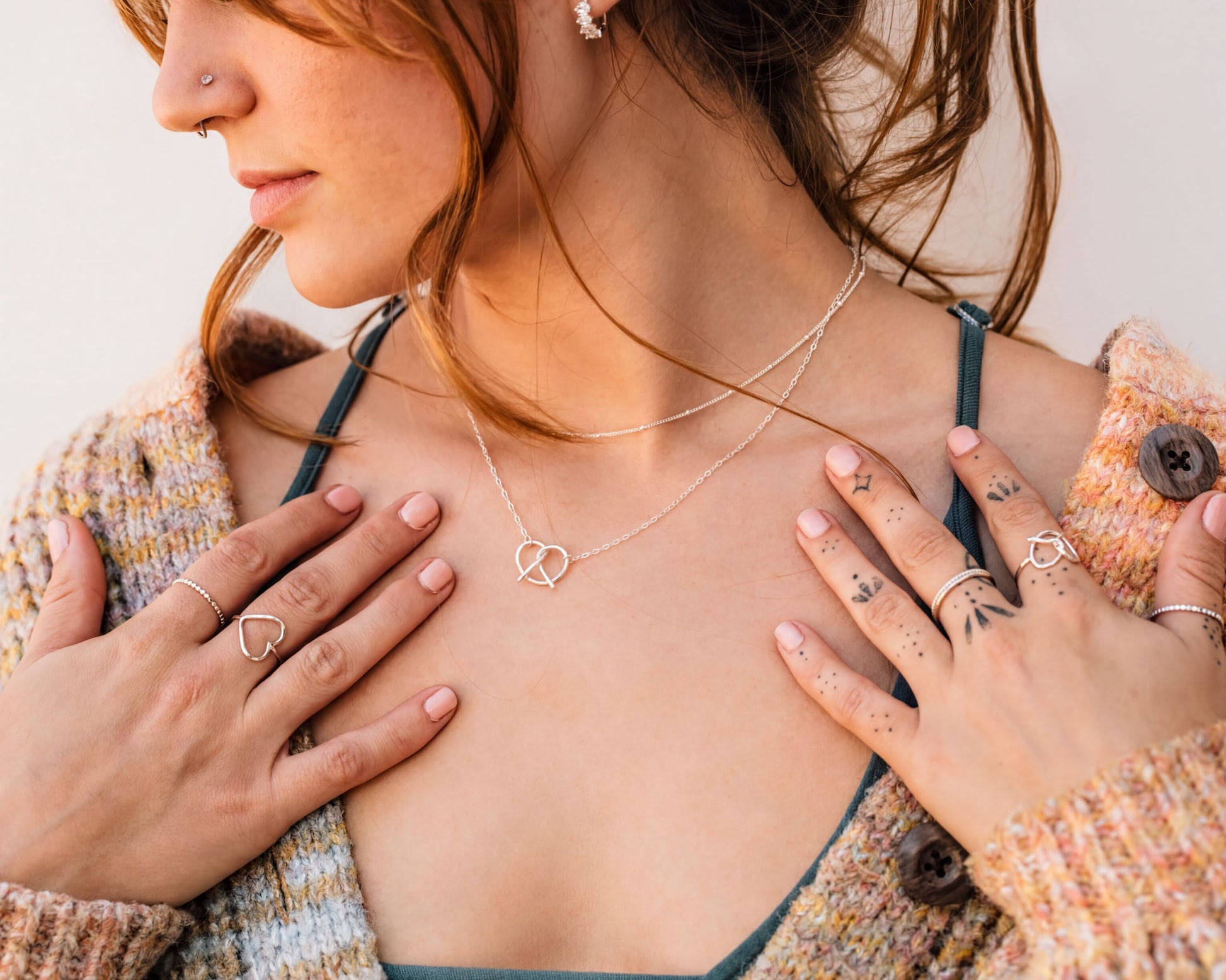 A close-up of a woman's face wearing a necklace and rings, with her hands placed on her chest.