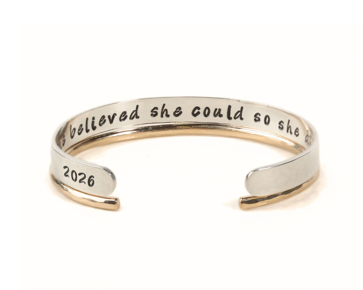 A silver and gold bracelet with the text "Believing she could so she" engraved on it.