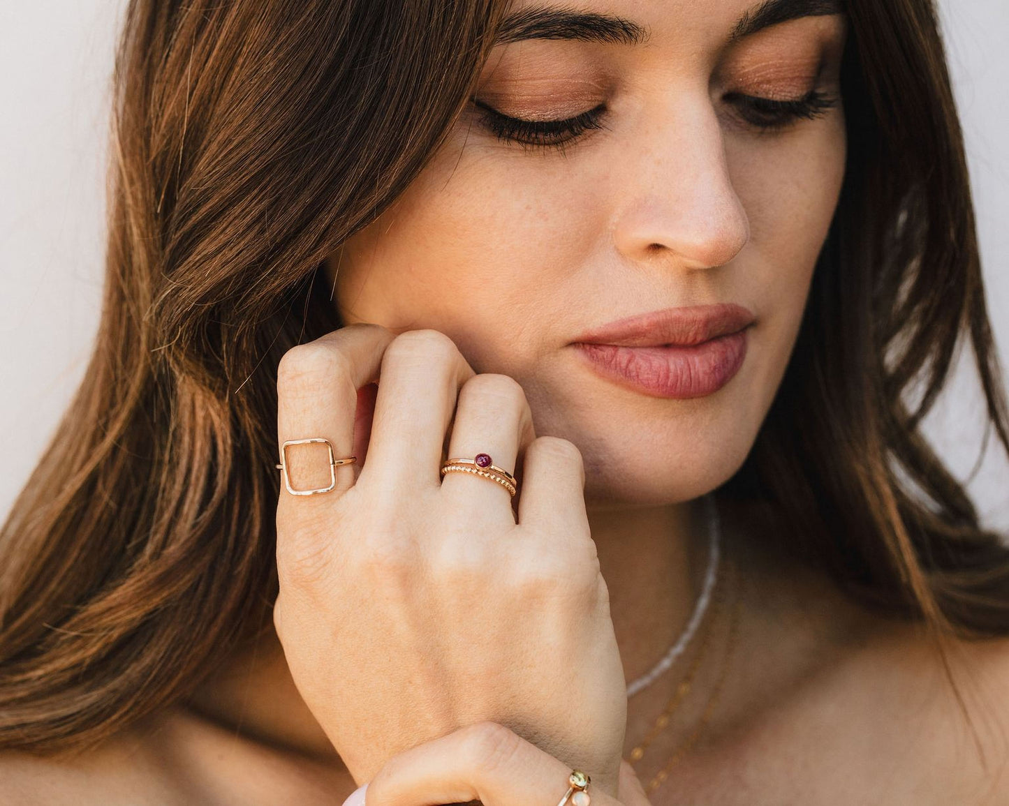 A close-up of a woman's face, with her eyes closed and her hand resting on her chin. She is wearing multiple rings on her fingers, including a ring with a gemstone and another ring with a geometric design.