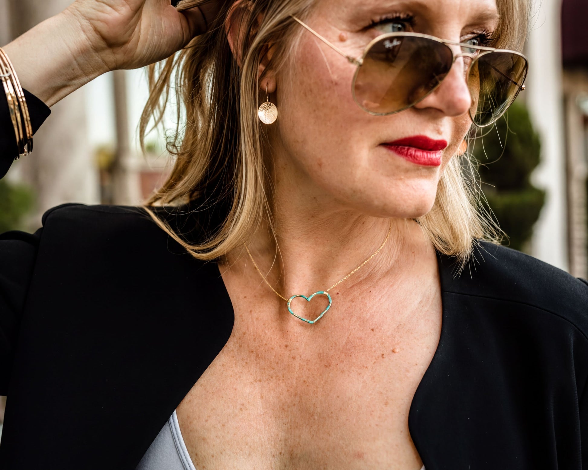 A woman wearing a black jacket, sunglasses, and a heart-shaped necklace stands outdoors.