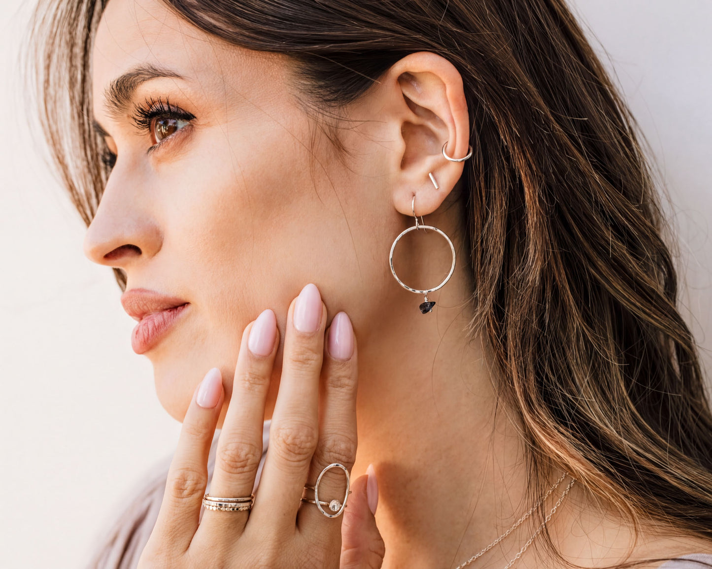 A woman with long brown hair wearing multiple rings and earrings, including a large hoop earring and a ring with a small gemstone.