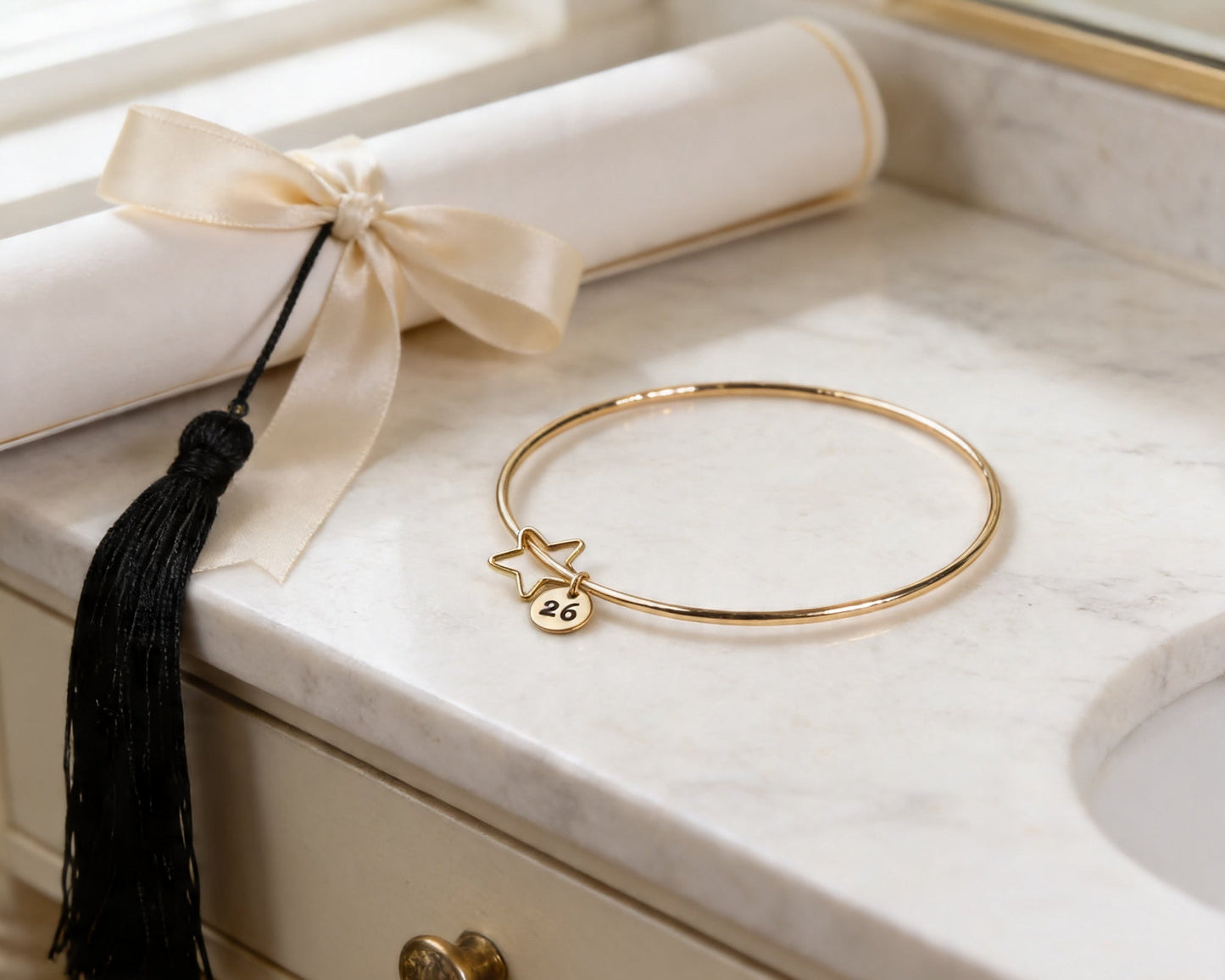 a gold bangle bracelet with a charm and a white tassel on a marble countertop.
