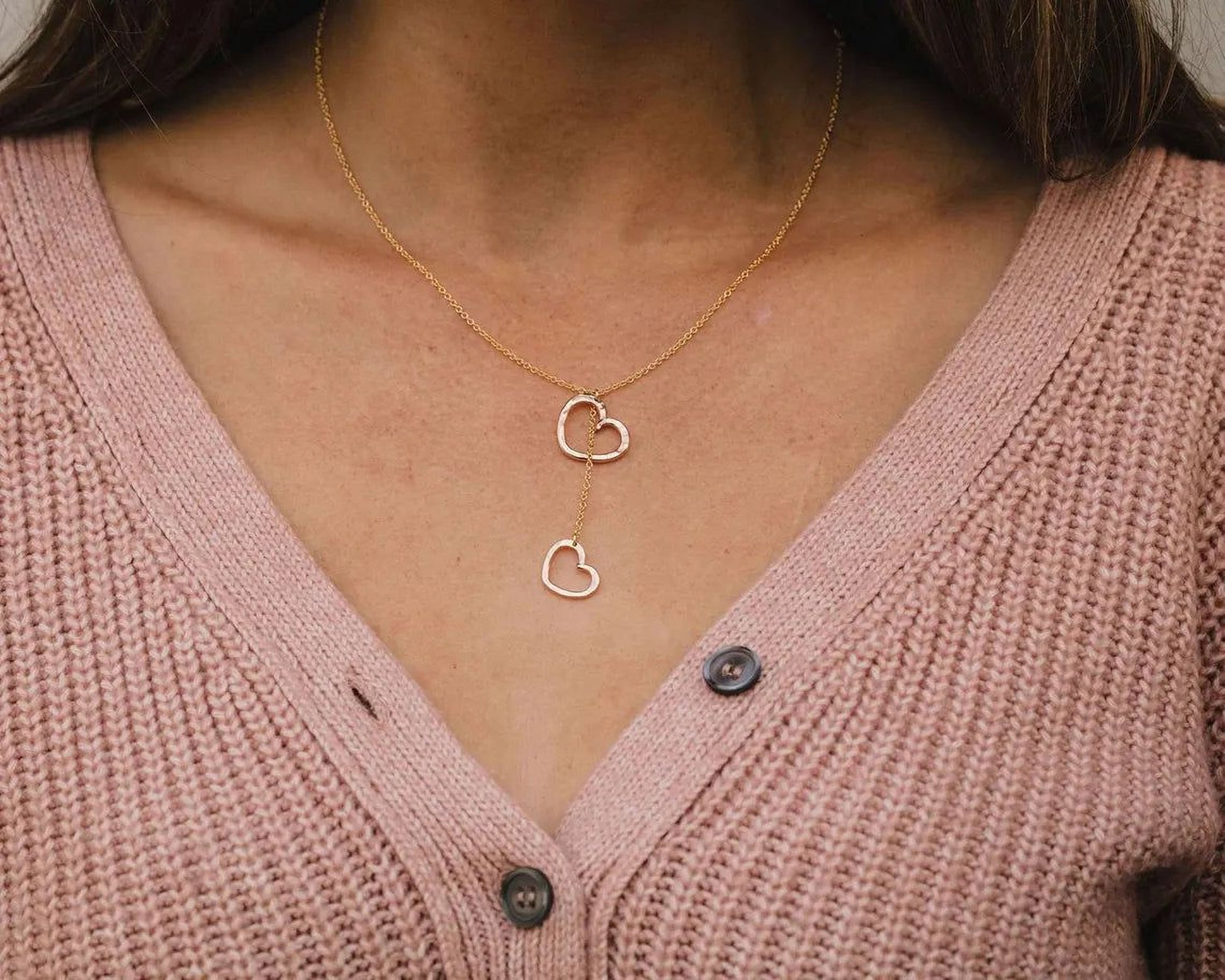 A close-up view of a woman wearing a pink cardigan and a gold necklace with two heart-shaped pendants.