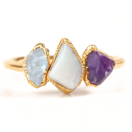 A gold ring with three gemstones: a white opal, a purple amethyst, and a blue sapphire.