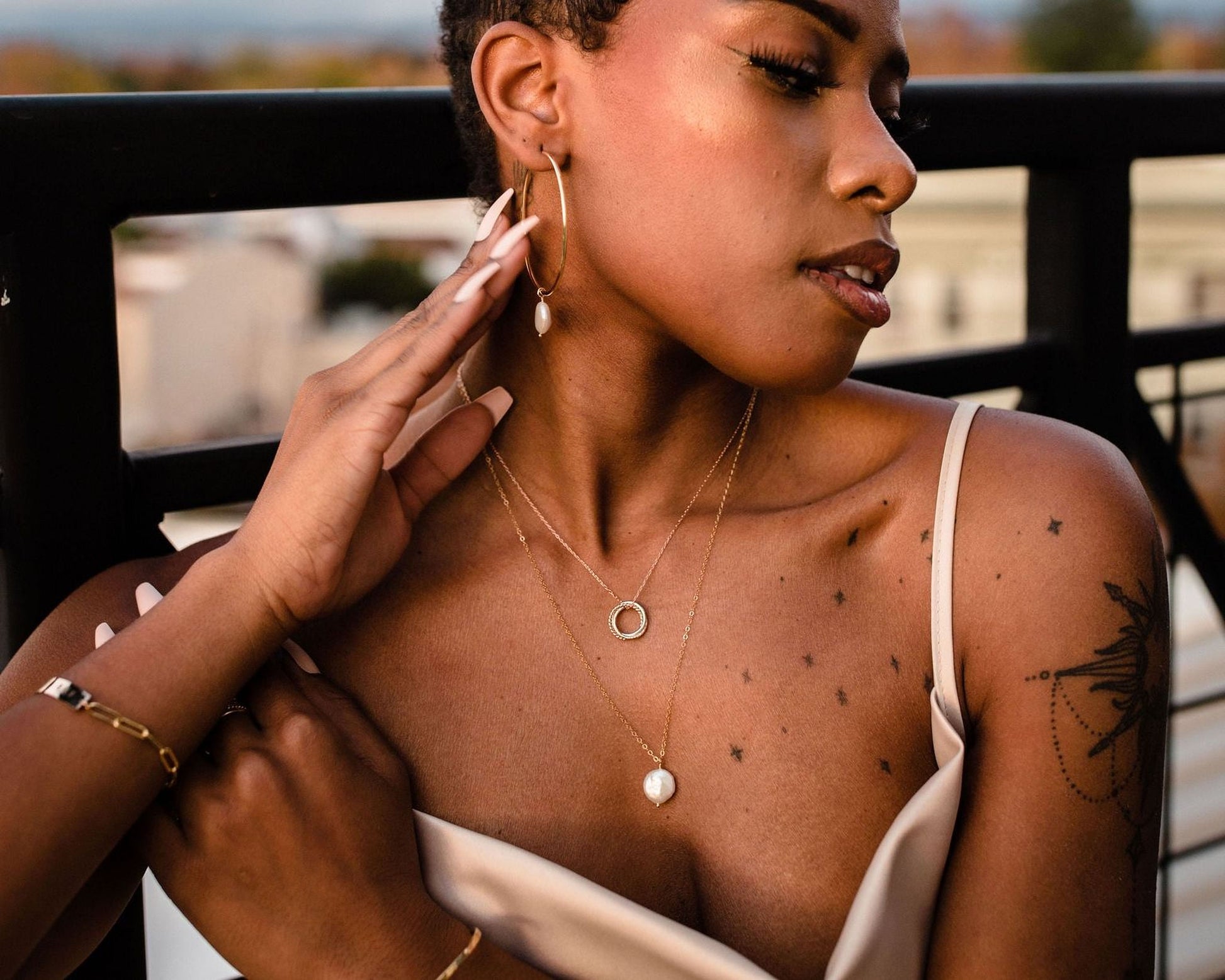 A woman wearing a white dress and multiple necklaces is shown in a close-up shot, with her hand resting on her chin.