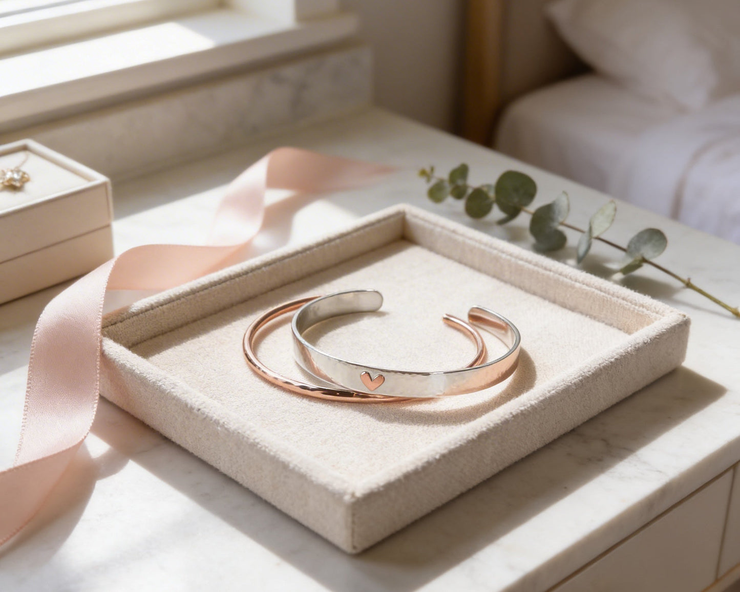 A pair of copper-colored bracelets is displayed on a beige tray, accompanied by a pink ribbon and a sprig of greenery, all placed on a white marble surface.