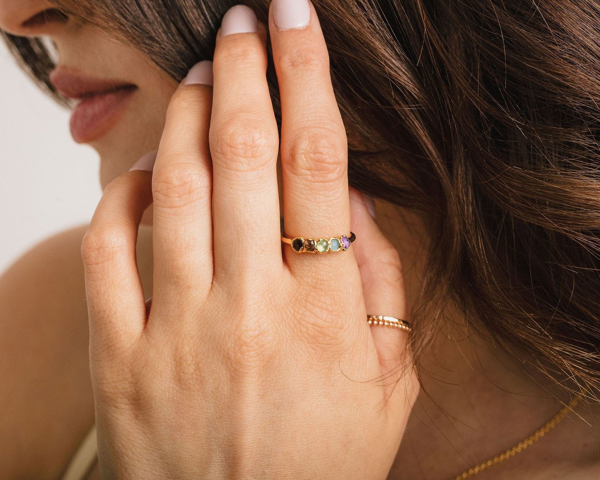 A close-up of a person's hand wearing a ring with a colorful gemstone band, partially covering their face.