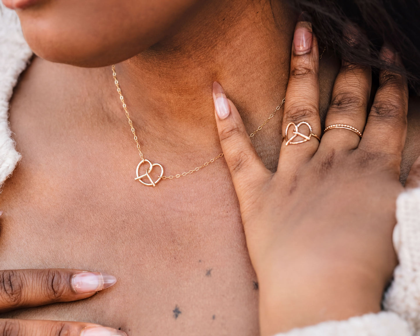 A close-up of a person's neck and chest area, featuring a delicate gold necklace with a heart-shaped pendant and a matching heart-shaped ring.