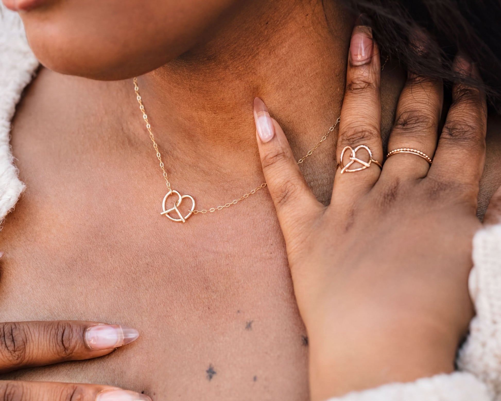 A close-up of a person's neck and chest area, featuring a delicate gold necklace with a heart-shaped pendant and a matching heart-shaped ring.