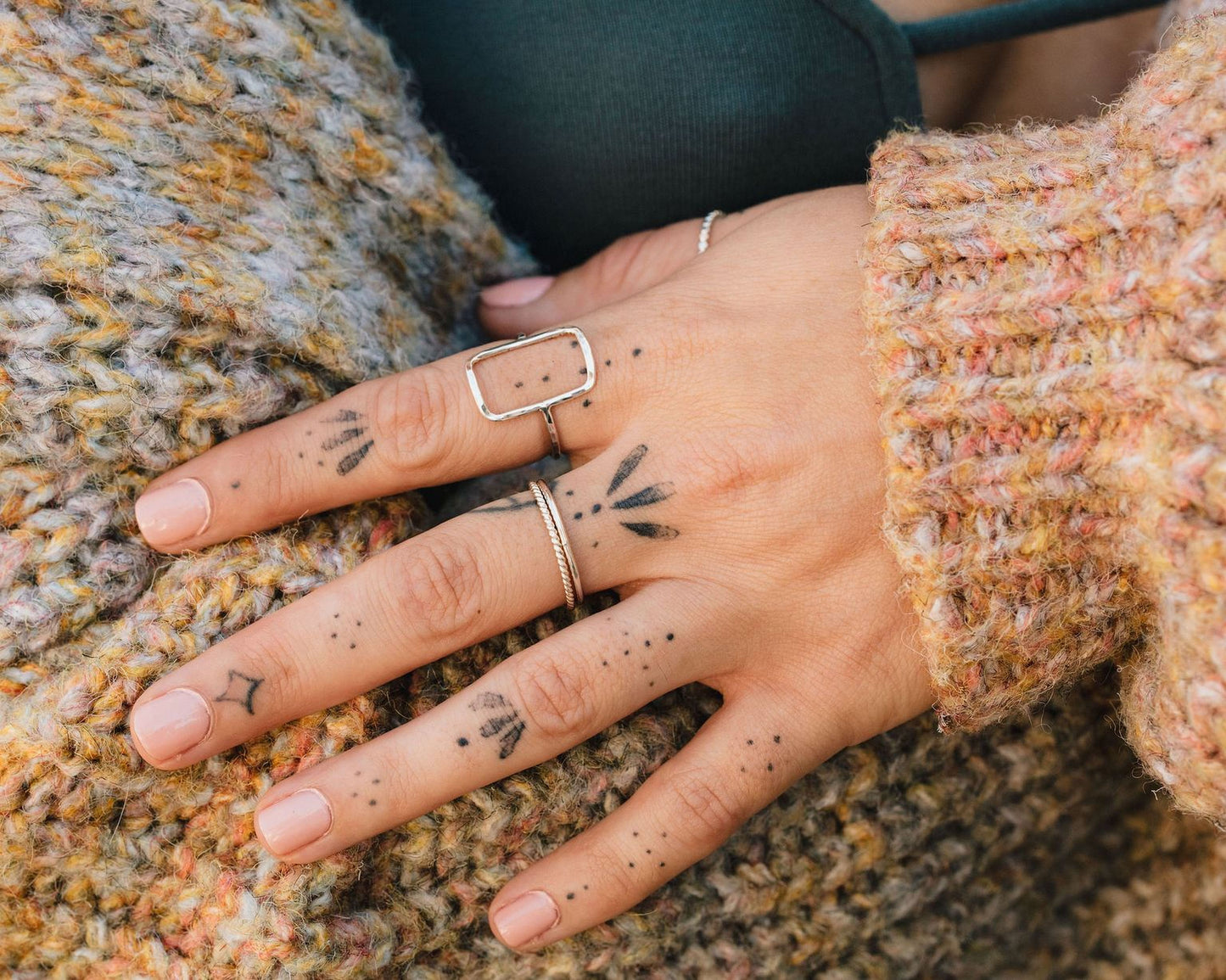 A close-up image of a person's hand adorned with various tattoos on the fingers and palm area, wearing a cozy sweater.