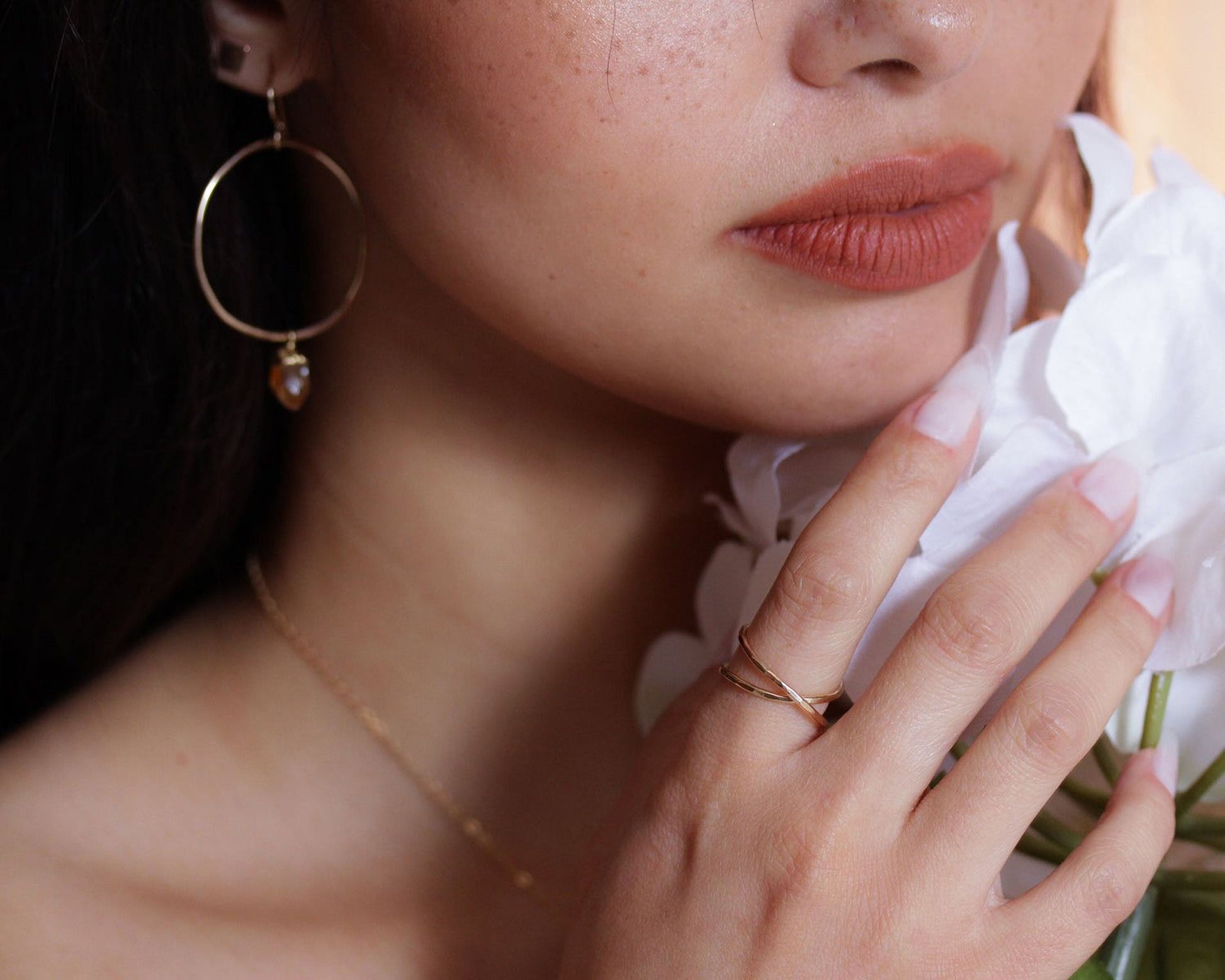 A close-up of a woman's face, with her hand holding a white flower. She is wearing a gold necklace and earrings, and has red lipstick on her lips.