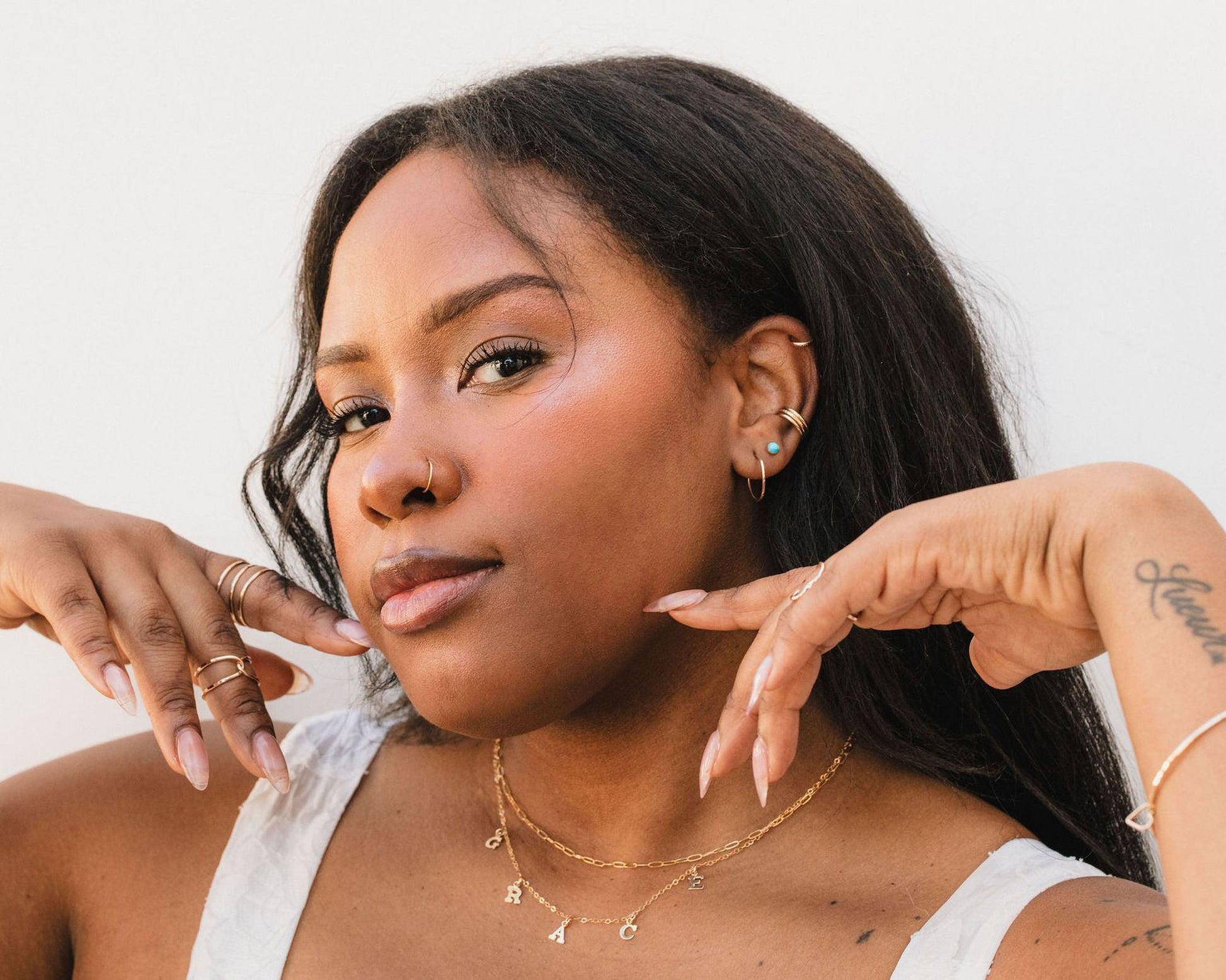A close-up portrait of a woman with dark hair, wearing a white top and jewelry, including multiple rings and earrings.