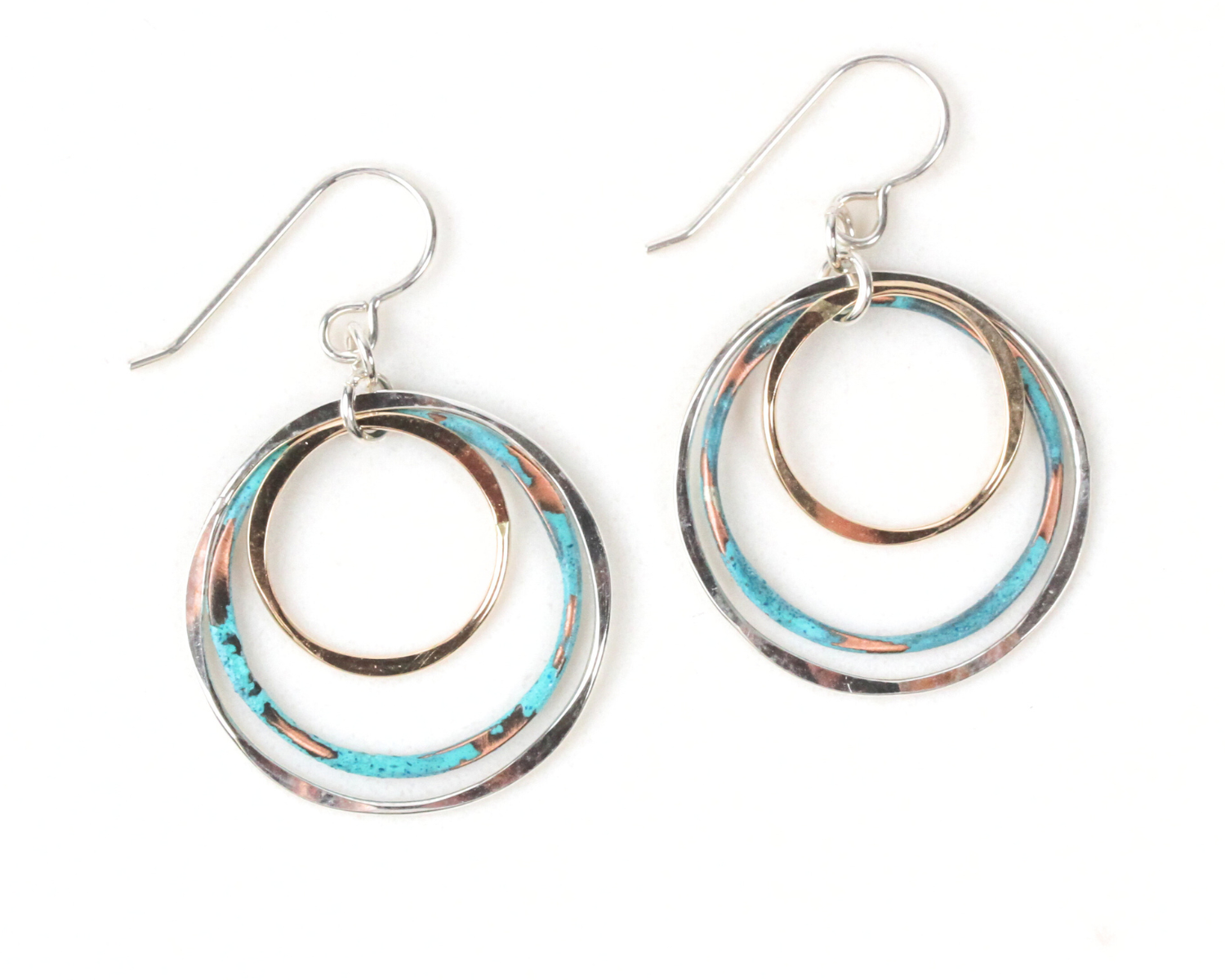 When color, quality, and artistry align, magic happens. These vibrant mixed metal earrings combine lustrous fine metals with a pop of rustic mint color and spin it all together in a delightful dance. Organically shaped hoops make up this three circle earring design. Each is meticulously handcrafted. The largest .925 Sterling Silver hoop measures approximately 1”. Mint patina over Copper and 14 Karat Gold Filled hoops gleam within. Our .925 Sterling Silver ear wires complete these delicate three circle earri