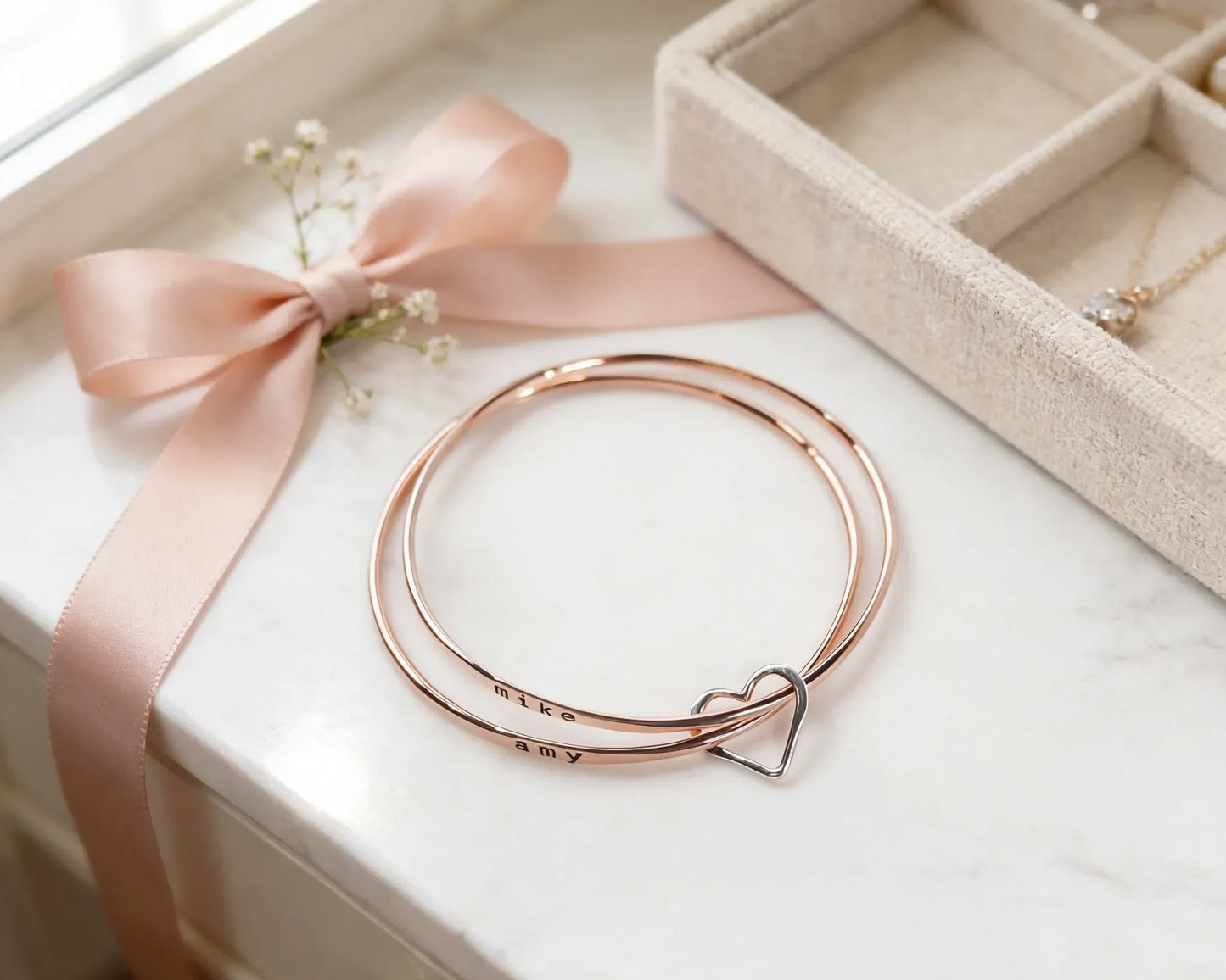a white marble surface with a pair of gold-colored bracelets, a pink ribbon, and a small potted plant.