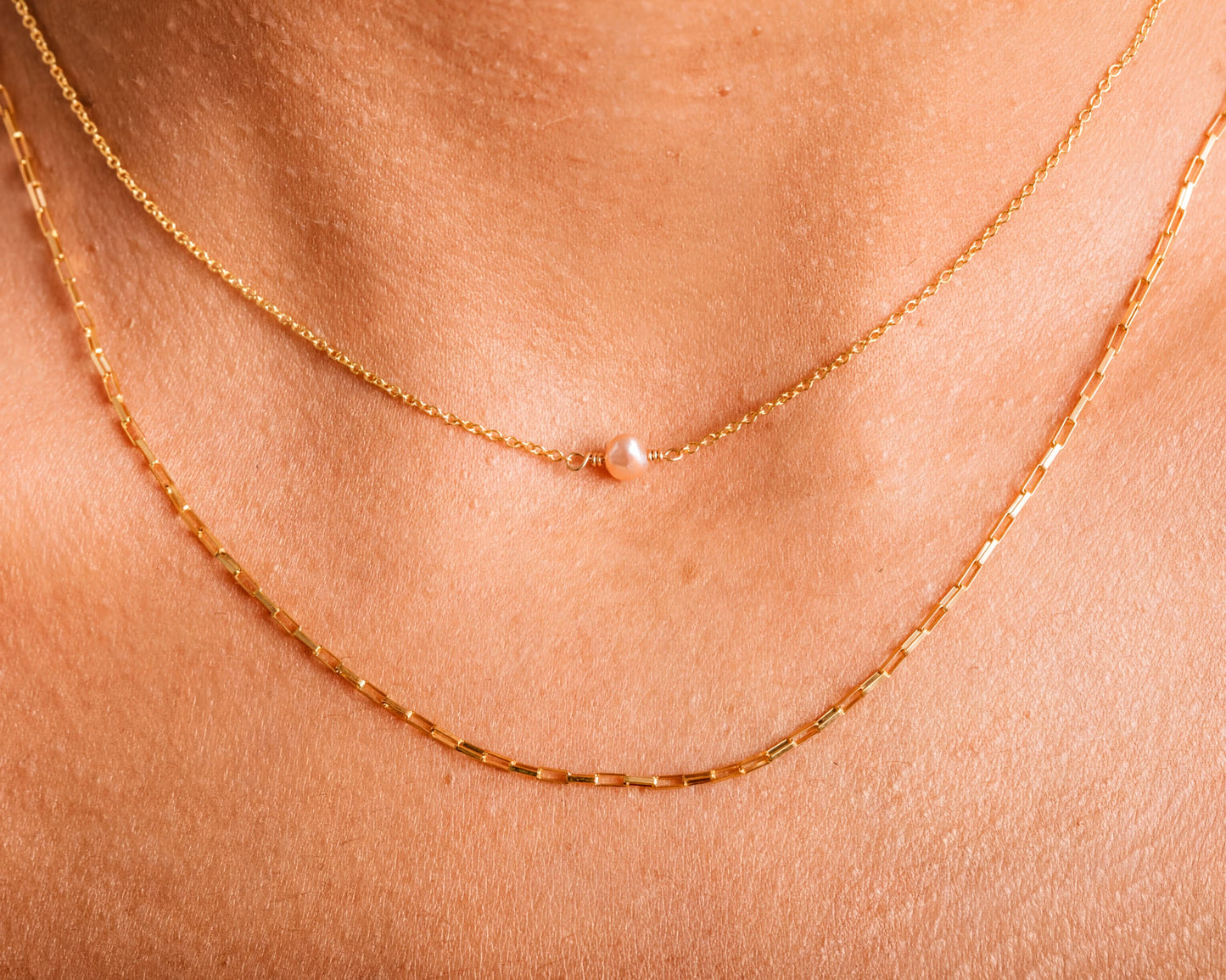 A close-up view of a person's neck adorned with two gold necklaces. The necklace on the left is a delicate chain with a single pearl pendant, while the necklace on the right is a longer chain with multiple pearls.