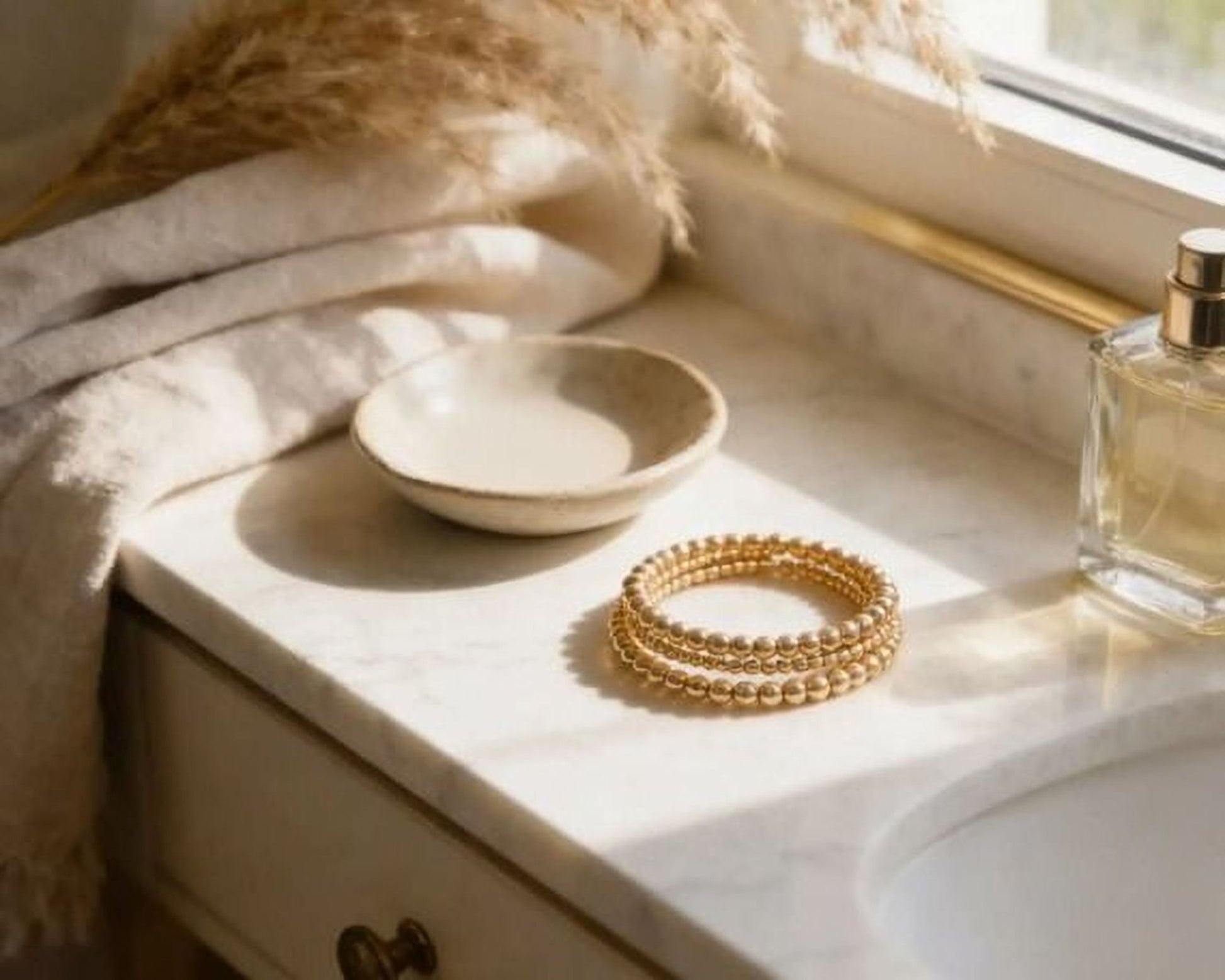 a marble countertop with a small white bowl, a gold bracelet, and a perfume bottle.
